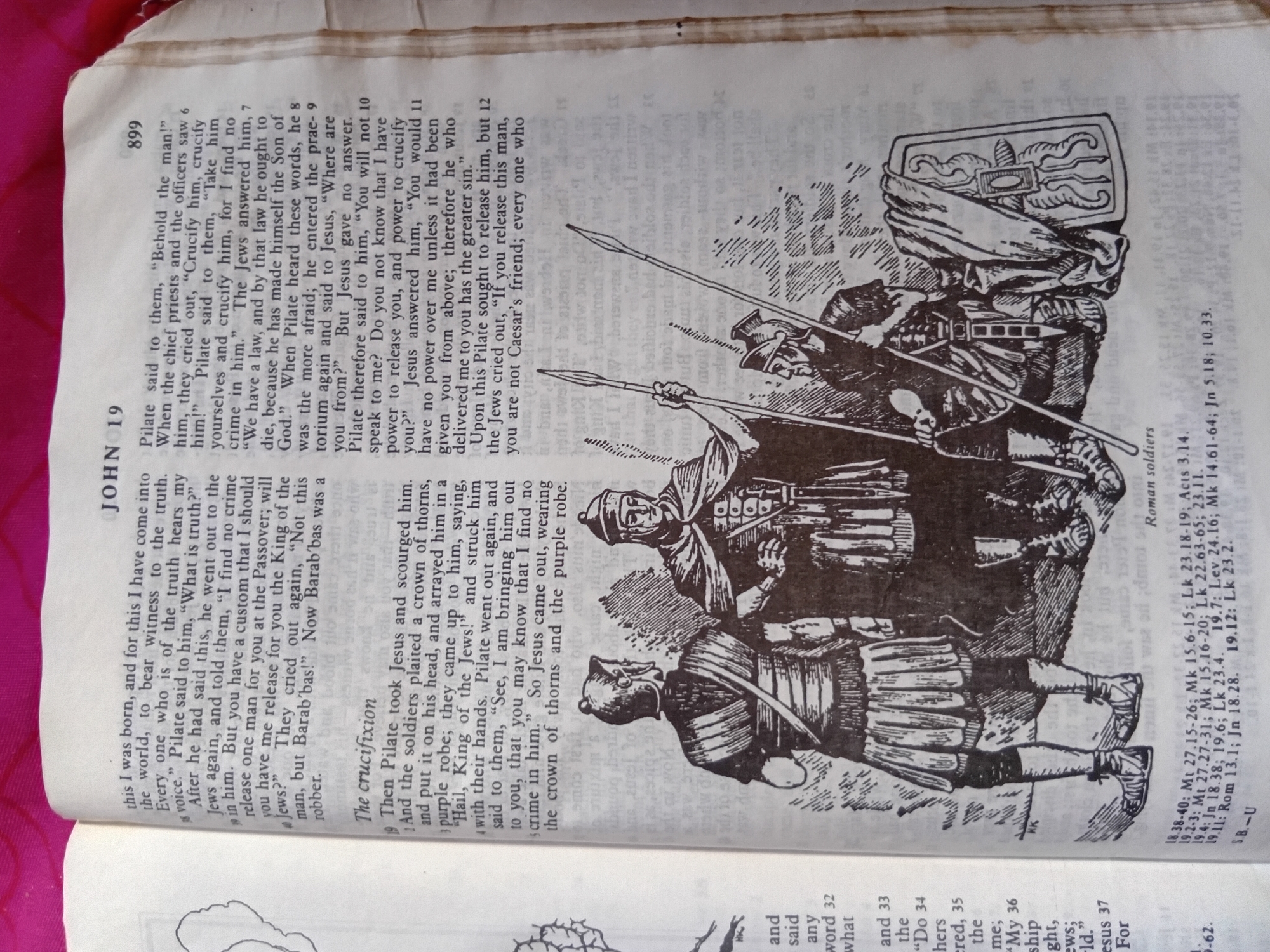 The image shows a page from a book, likely a Bible, with text and an illustration. The text is in English and features a chapter heading "JOHN 19". The illustration depicts several Roman soldiers in historical attire, including armor, helmets, and shields. One soldier is shown holding a shield with a decorative pattern. The soldiers appear to be engaged in a scene of confrontation or procession, with spears or javelins. The page is partially covered by a pink fabric. The text to the right of the illustration includes verse numbers and references such as "Mk 15.6-15", "Lk 23.18-19", "Jn 19.1-3", "Mt 27.27-31", "Lk 22.63-65", "Acts 4.27", "Jn 18.38", "Mk 14.61-64", "Jn 10.33", "Lev 24.16", "Mk 14.61-64", "Jn 18.28", "Lk 23.2". The city of Nkpor, Nigeria is not depicted or mentioned in the image.