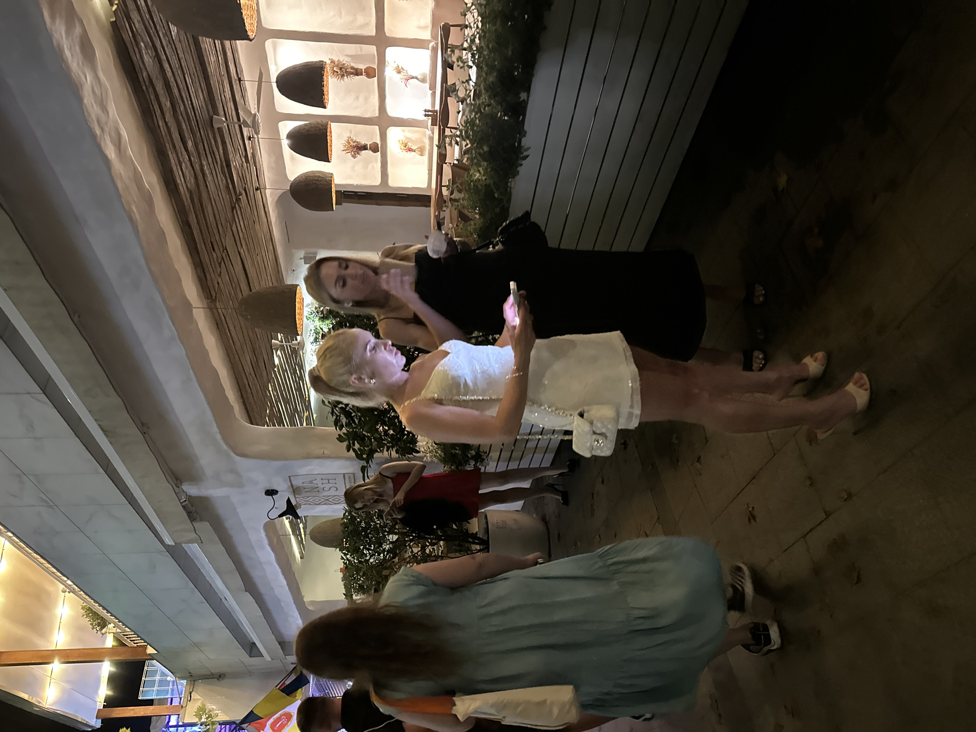 The image shows an outdoor scene at night, likely in Odesa, Ukraine, given the context. Four individuals are visible. On the left, a person with long dark hair and wearing a teal dress is walking away from the camera, carrying a white tote bag with an orange strap. To their right, a woman in a red dress with black high heels is standing and looking down at her phone. Next to her, a woman in a short, sparkly white dress with a white handbag is looking at her phone. To the right of her, a woman in a long black dress is standing, holding a white object, and looking down. In the background, there is a building with an awning, decorative lighting, and a sign that reads "NASH". There are also plants, tables, and chairs visible. The ground is paved.