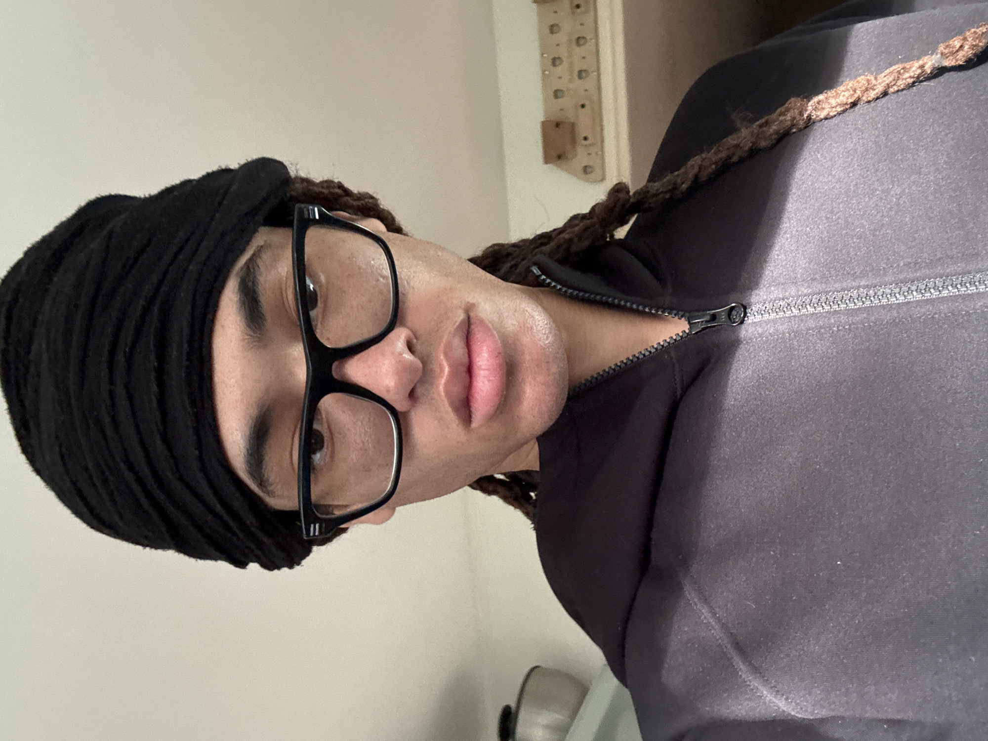 A close-up, portrait-style image, taken indoors and rotated 90 degrees clockwise, features an individual looking directly at the camera. The person has dark skin, prominent lips, and visible eyebrows. They are wearing black-framed glasses that obscure part of their eyes. Their hair, styled in dark brown dreadlocks or braids, is partially covered by a black headwrap or durag. Several braids are visible around the neck and shoulder area.
The individual is dressed in a dark-colored, possibly black or charcoal, long-sleeved jacket with a visible silver zipper running down the front. The fabric of the jacket appears soft and slightly textured. The person's expression is neutral, with a slight pout to the lips.
Natural light illuminates the scene, casting highlights on the face, particularly the forehead and lips, and creating soft shadows. The background is simple, featuring a light-colored wall. On the right side, a light wooden panel with evenly spaced holes and metal pegs is partially visible, suggesting a utility board or decorative item. The overall setting appears to be a well-lit indoor space, possibly a room or hallway. No text is visible in the image. The image is contextualized as being taken in Les Ulis, France.