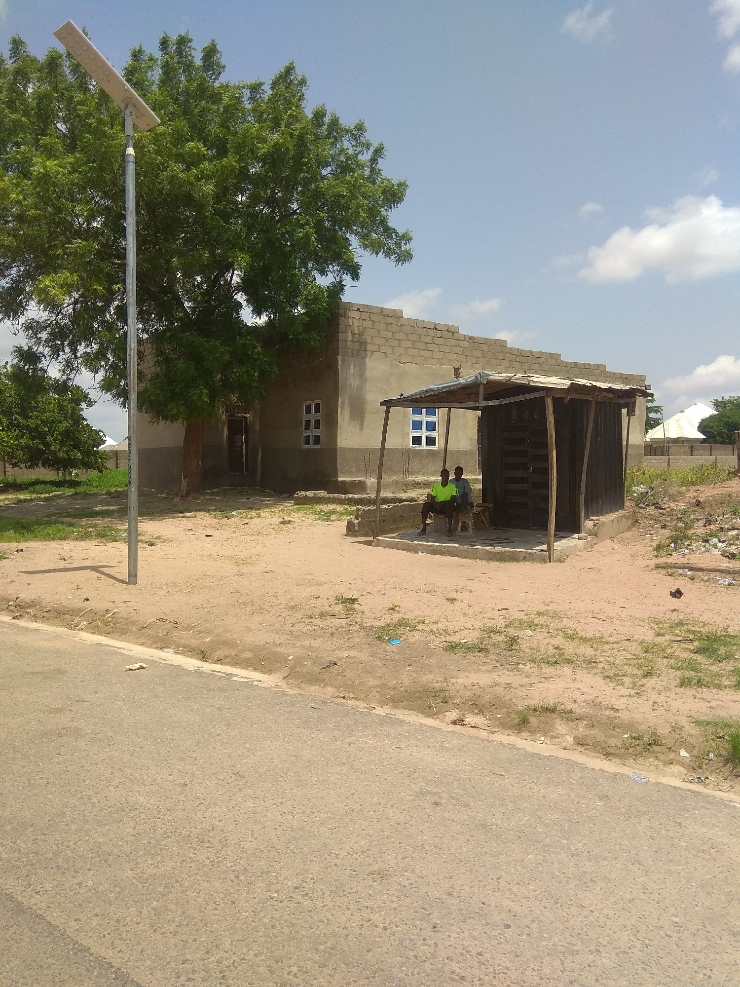 This image captures an outdoor scene in Jalingo, Nigeria, on a bright, sunny day with a few scattered clouds. The foreground features a paved road with a sandy shoulder, littered with some debris.
The main subjects are two individuals seated under a rustic, open-sided shelter constructed with wood and corrugated metal roofing. The shelter is adjacent to a partially constructed building made of concrete blocks. One person is wearing a bright green t-shirt and is sitting on a wooden bench, while the other, wearing a blue shirt, is seated on what appears to be a stool. They appear to be resting or waiting.
A tall, modern solar-powered street light stands prominently to the left of the frame, its panel angled towards the sun. Behind the shelter and the partially built structure, a large tree with dense green foliage dominates the middle ground, casting a shadow on the ground. Further in the background, more trees and buildings can be seen, including a white-roofed structure. The overall setting suggests a semi-rural or developing area. The time of day is clearly daytime, indicated by the strong sunlight and the clear sky. No visible text is present in the image.