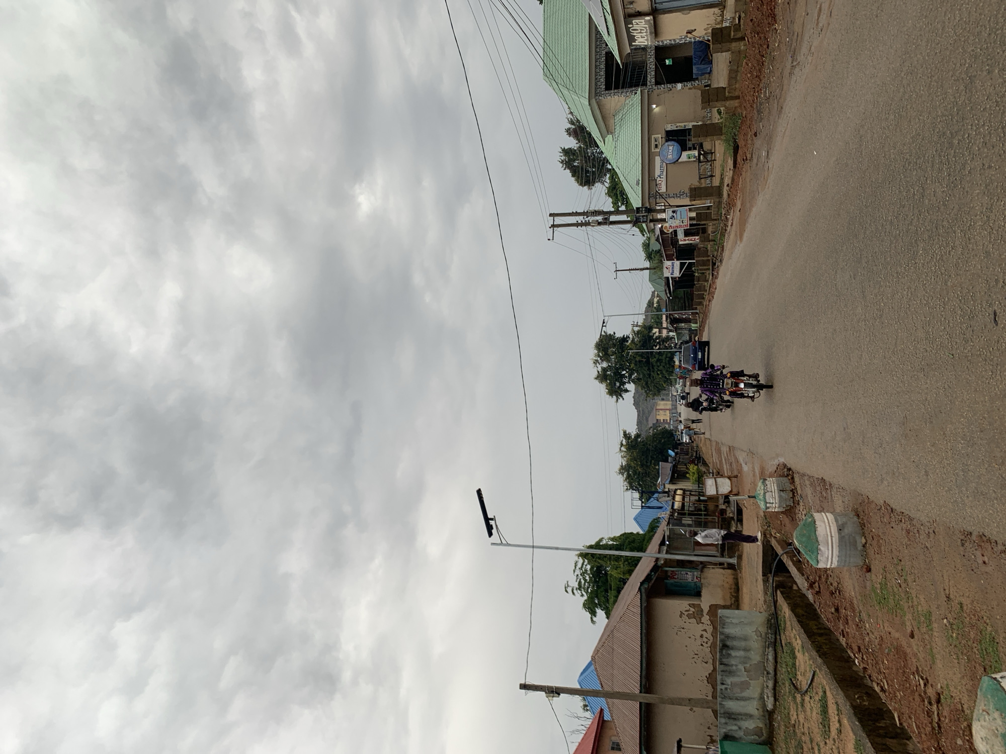 The image shows a street scene in the New Stadium area of Nigeria. The sky is overcast with grey clouds. A paved road runs through the scene, with buildings lining both sides. Several motorcycles are traveling on the road, with multiple occupants on each. There are utility poles with wires visible above the buildings and road. The buildings appear to be a mix of residential and commercial structures. There is some vegetation visible, including trees and bushes. Along the side of the road are what appear to be painted cement or concrete blocks. A street light stands on a pole.
