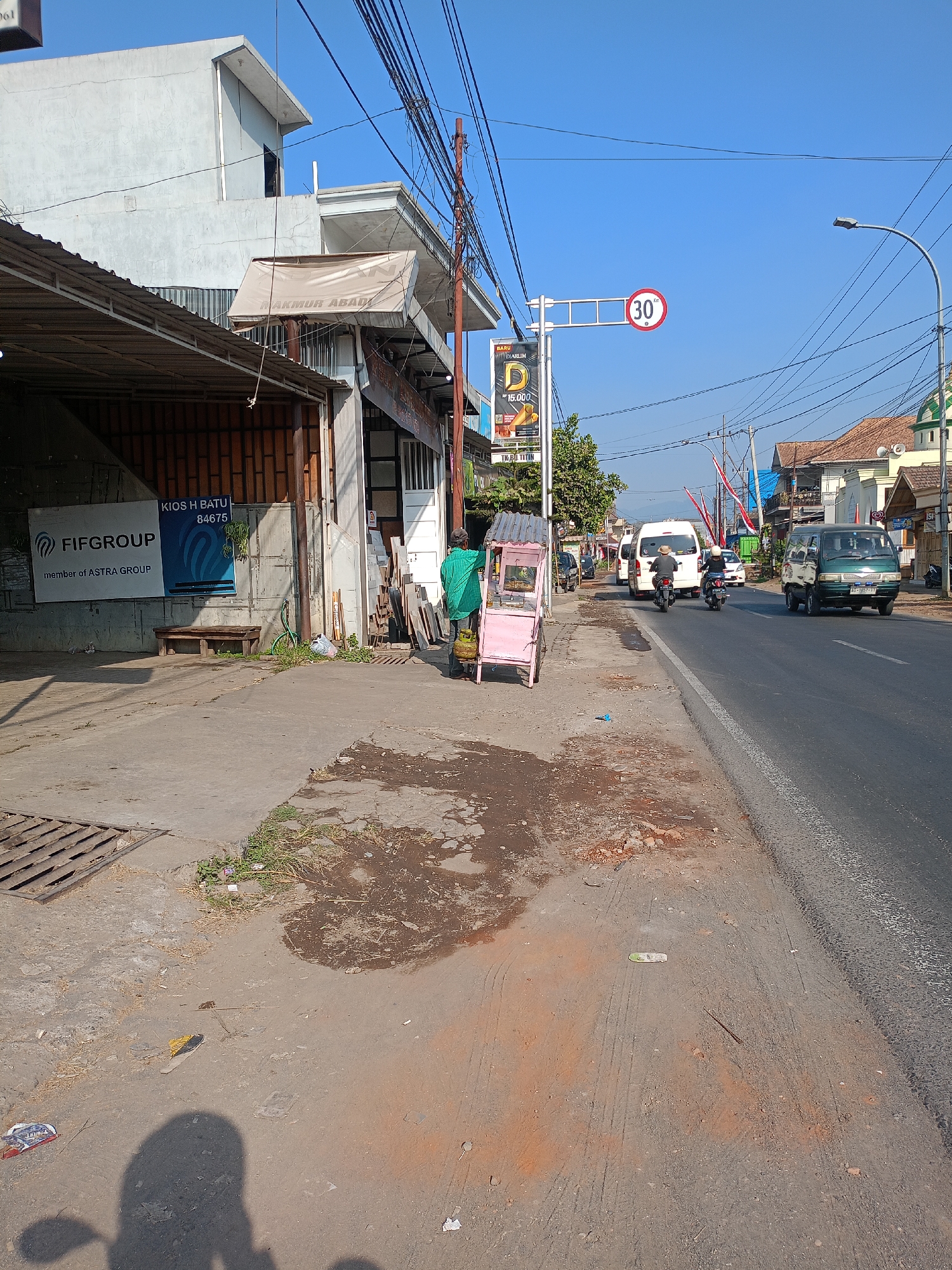 The image shows a street scene in Batu, Indonesia, with a focus on the side of the road and adjacent buildings. On the left, a storefront with a corrugated metal roof displays a sign for "FIFGROUP member of ASTRA GROUP" and "KIOS H BATU 84675". A wooden bench is positioned in front of the storefront. Further down the street, a pink, three-wheeled cart with a canopy is parked on the sidewalk.
The road itself is paved and has visible lane markings. Several vehicles are present, including two motorcycles, a white van, and a green van. Traffic appears to be moderate. Overhead, a complex network of power lines is strung across the sky. A speed limit sign indicating "30" km/h is visible, as is a street light. The sky is clear and blue. The ground in front of the buildings is a mix of concrete and dirt, with some patches of dry grass. Debris and a shadow of a person on a motorcycle are also present on the ground. In the background, more buildings and trees are visible, with a hint of distant mountains. The overall impression is of a typical urban street in a warm climate.
