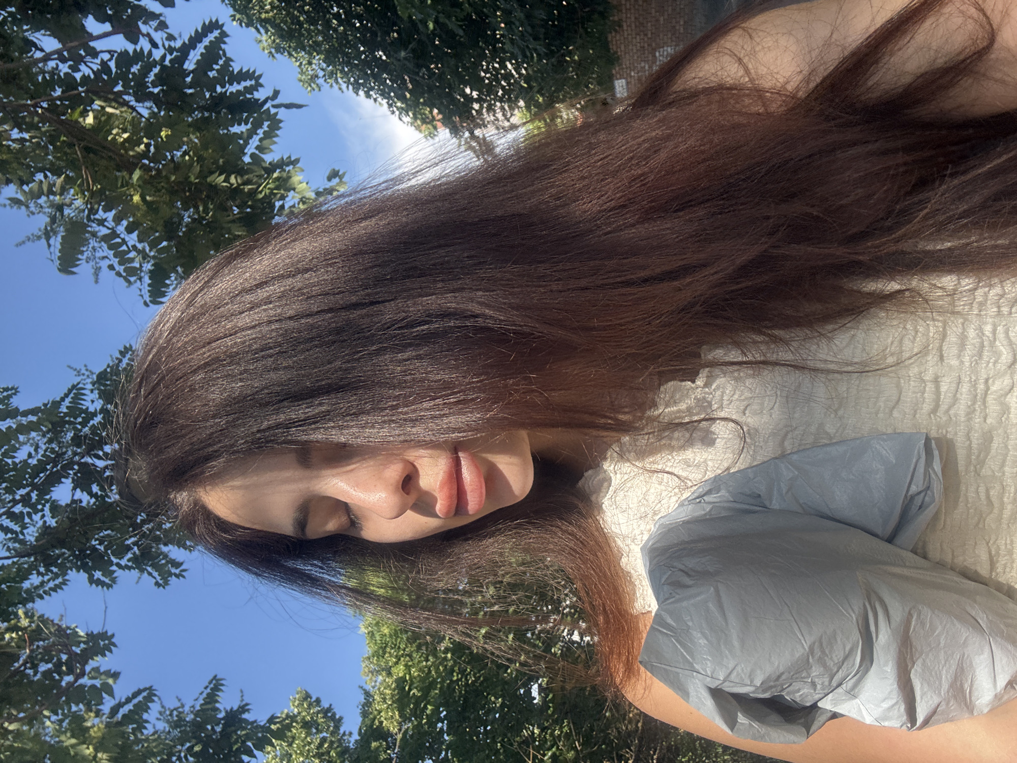 The image, oriented sideways, presents a close-up view of a person outdoors on a sunny day. The subject has long, dark brown hair that cascades across the frame, partially obscuring their face. Their skin is visible, and their eyes appear closed or are looking downwards. Prominent, full lips are visible. They are wearing a light-colored, possibly off-white, textured top with smocking.
In the lower right portion of the image, a crinkled, grey, almost silvery-blue object or fabric is visible, resting on what appears to be the person's arm or torso.
The background consists of a clear, bright blue sky and lush green tree foliage, with sunlight filtering through the leaves, creating bright highlights and dappled shadows. The trees suggest an outdoor setting, potentially a park or garden in Plovdiv, Bulgaria. The overall impression is one of a serene and relaxed moment. No text is visible in the image.