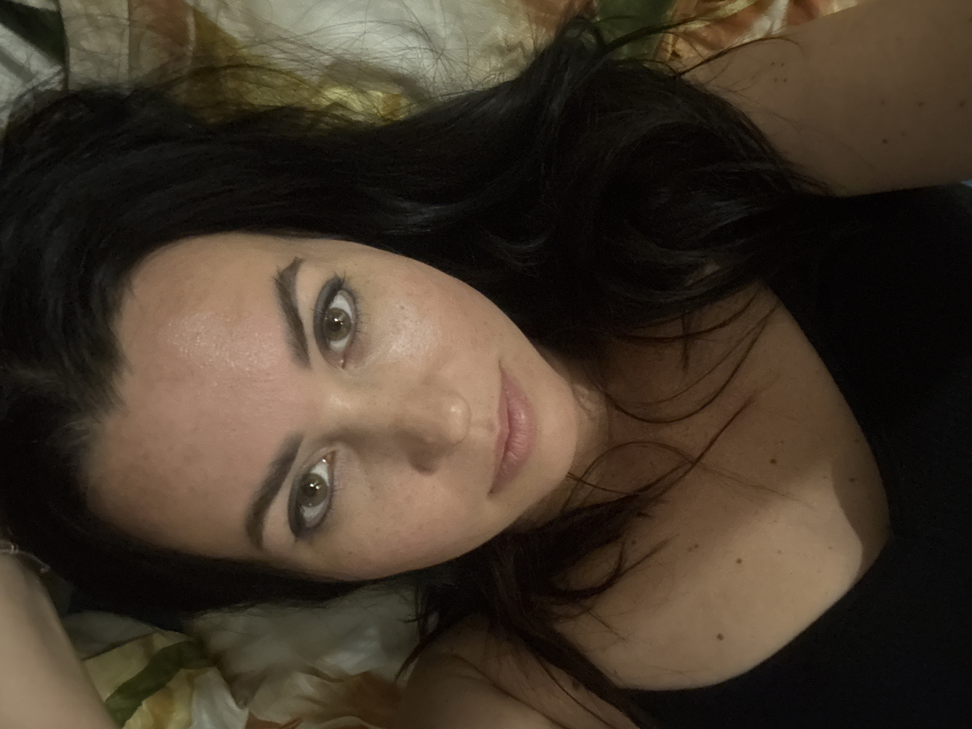 A woman with dark hair is lying on a patterned surface, possibly a bed. She is facing the camera and looking up. Her skin appears fair with freckles. She is wearing a dark, possibly black, top. There is no visible information to establish the location as Pattaya City, Thailand.