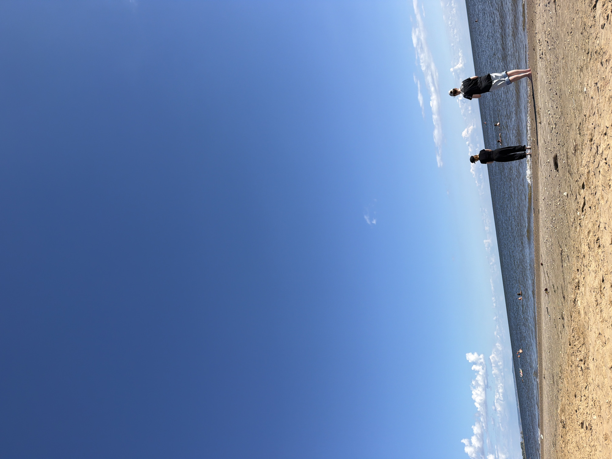 Two individuals stand on a sandy beach facing a calm body of water under a clear blue sky with scattered white clouds. The person on the left is wearing a black hat, black t-shirt, and black pants, with their back to the camera. The person on the right is wearing a black t-shirt and light blue shorts, with their back and right side visible. Both are looking out towards the water. In the distance, several other people are visible in the water, some swimming and others possibly on flotation devices. The sand on the beach is light brown and textured, with some small debris visible. The water is a deep blue, meeting the horizon where the sky fades into a lighter blue. The scene suggests a relaxed day at the beach.