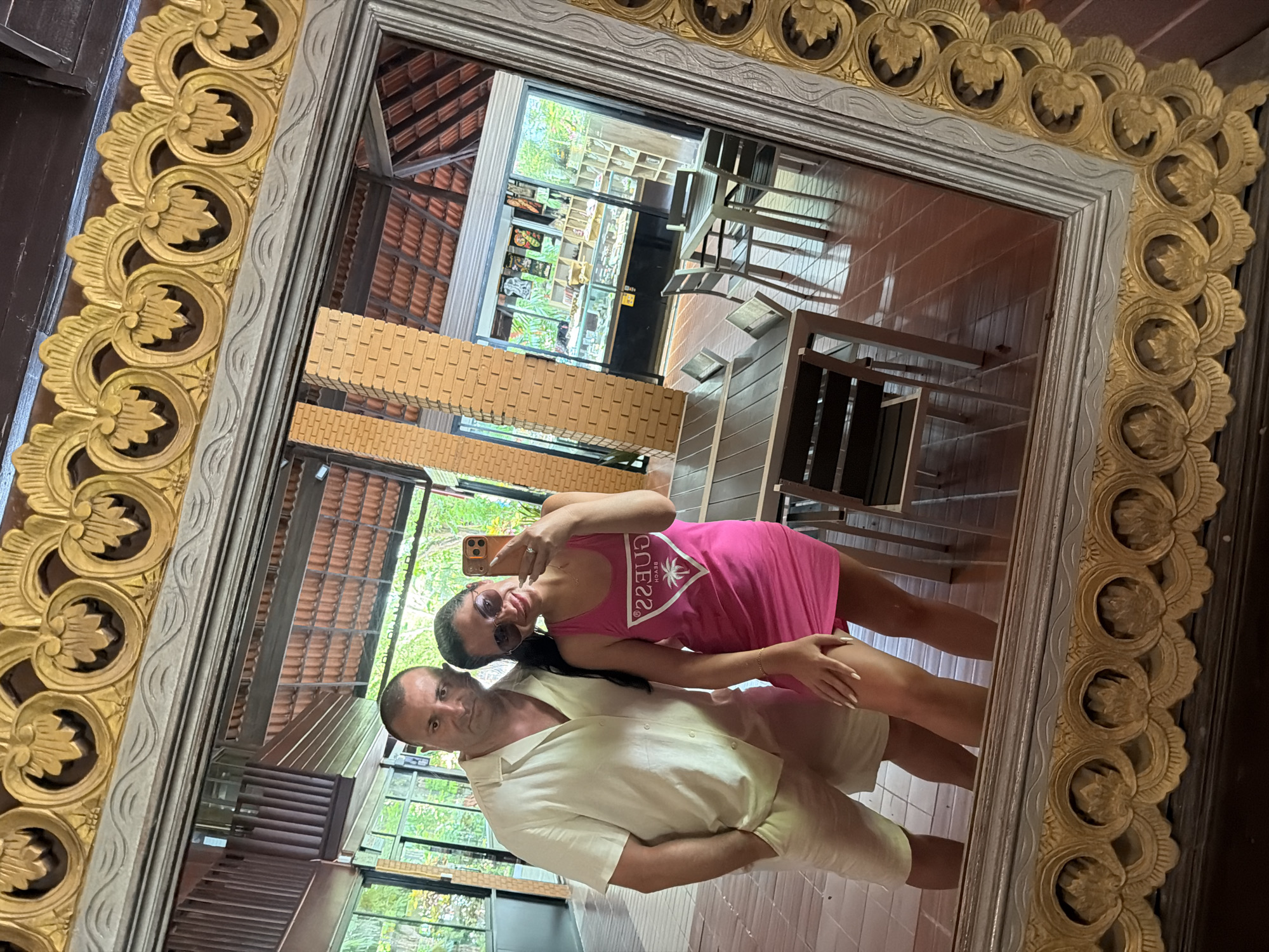A couple, a man and a woman, are taking a mirror selfie inside what appears to be a resort or restaurant in Ao Nang, Thailand. The man is on the left, wearing a light-colored short-sleeved button-down shirt and matching shorts. The woman is on the right, wearing a bright pink tank dress with a "Guess Beach" logo. She is holding her phone to take the photo. The background shows an open-air building with a tiled roof, brick pillars, and large windows revealing tropical foliage. Tables and chairs are visible in the reflection of the mirror, suggesting a dining area. The mirror frame is ornate and gilded, with intricate carvings. The overall impression is a casual, holiday snapshot in a tropical setting.