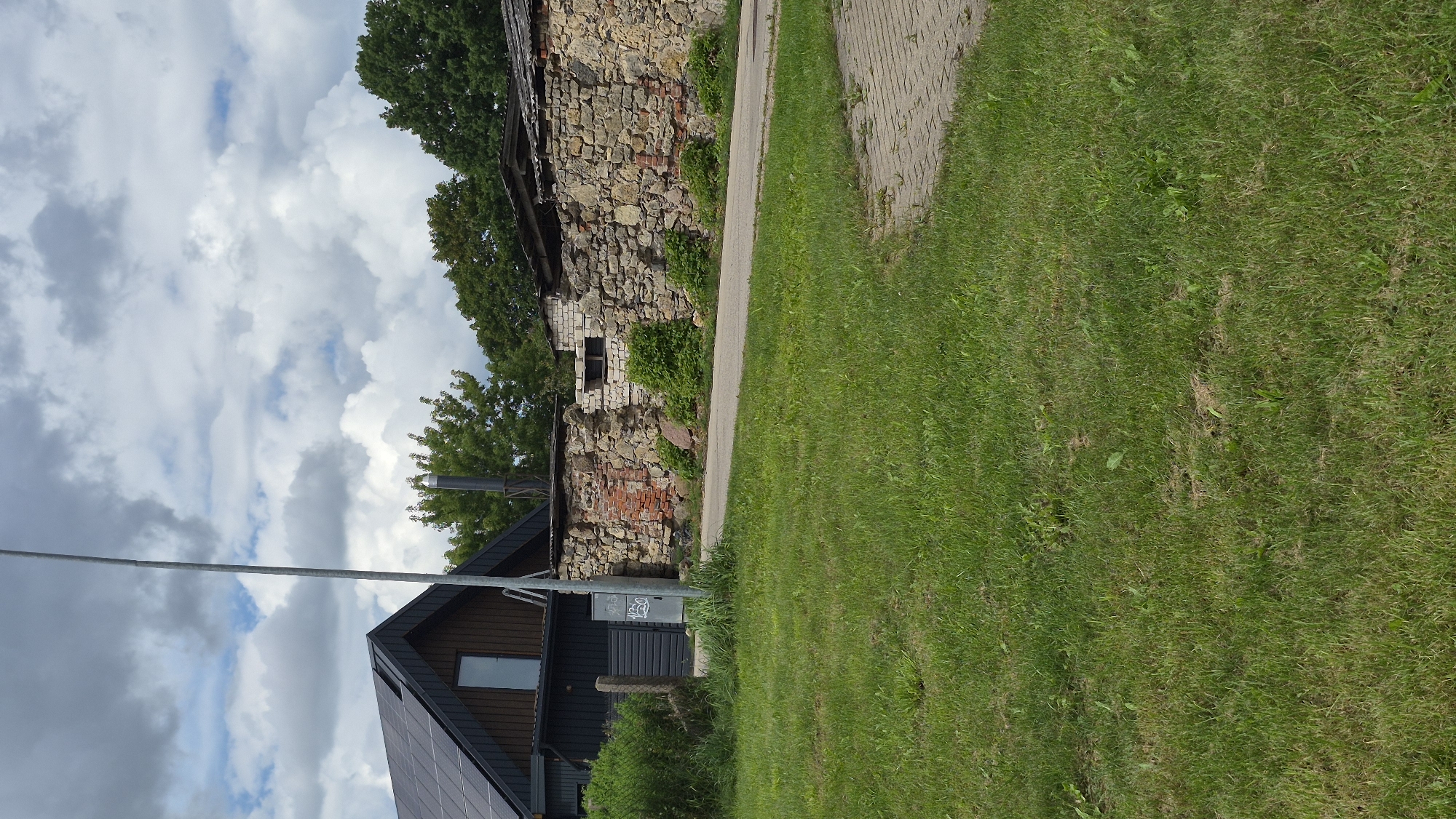 The image captures an outdoor scene in Jēkabpils, Latvia, featuring a rustic stone wall in disrepair, a paved pathway, and a grassy area. The stone wall, constructed from irregular stones and some exposed red brick, appears old and weathered. Several small green bushes are growing at the base of the wall. Behind the wall, glimpses of a building with a corrugated roof and what looks like part of another structure are visible. To the left, a dark-colored modern building with a dark roof, possibly covered in solar panels, is partially visible. A tall, slender metal pole stands in front of this building. The sky above is partly cloudy with a mix of white and gray clouds. The overall impression is one of a somewhat overgrown and aged rural or semi-rural setting.