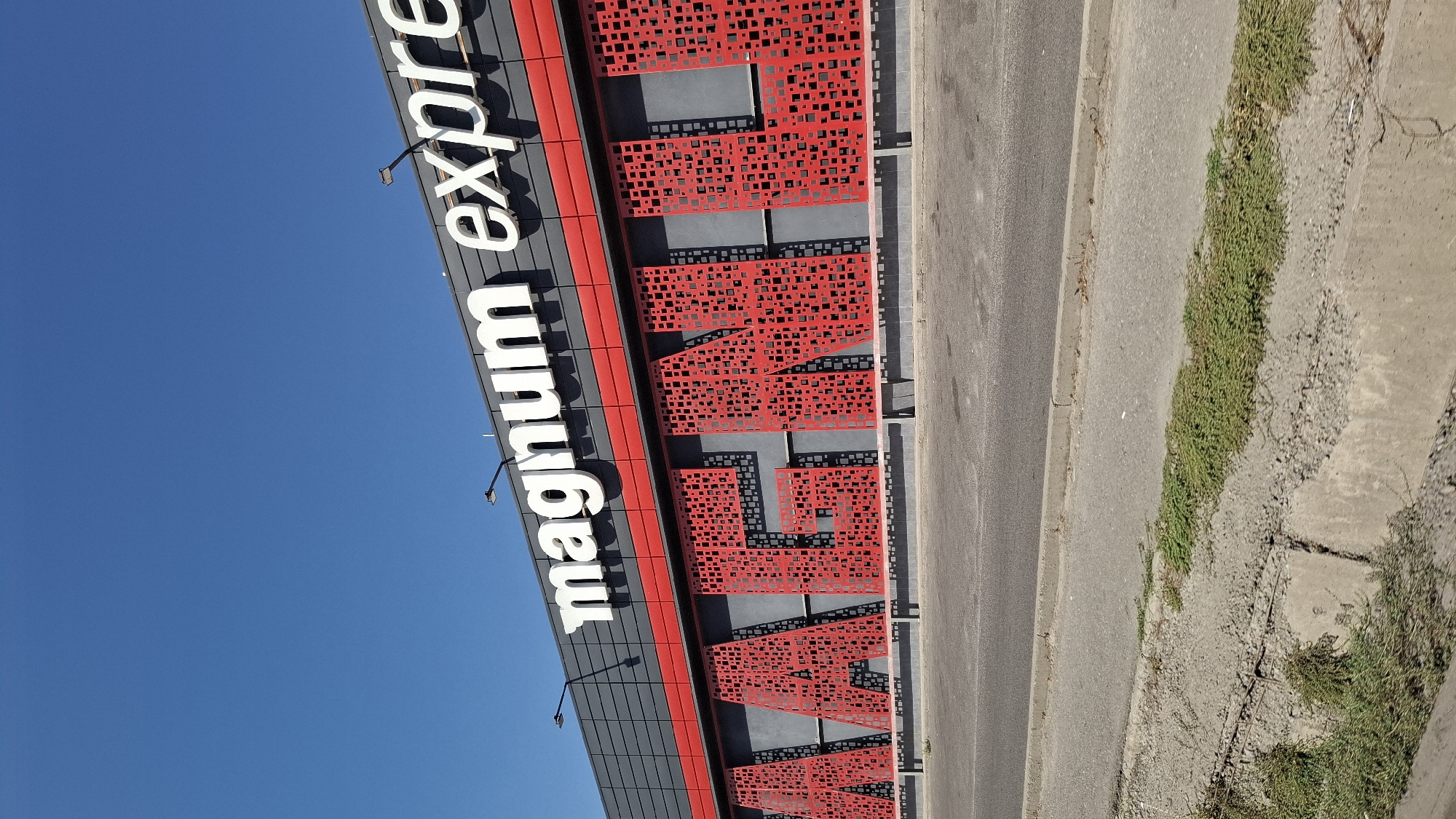 The image shows the exterior of a Magnum supermarket in Shymkent, Kazakhstan. The building has large white lettering on the top that reads "magnum express". Below this, large red decorative letters spell out "MAGNUM". The building is gray and red. In front of the building is a road and a sidewalk. The sky is clear and blue.