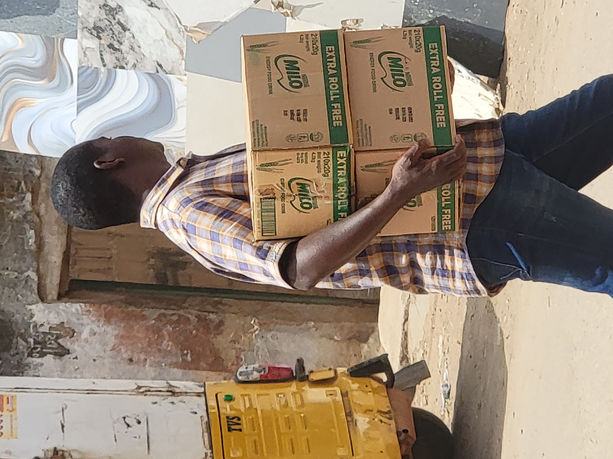 A man stands on a beige-colored dirt street in the city of Maiburiji, Nigeria. He is holding four brown cardboard boxes of Milo energy food drink with green "EXTRA ROLL FREE" labels. He is wearing a brown and white checkered shirt and dark blue jeans. Next to him, a yellow machine is partially visible, with "TVS" printed on it. The background features a weathered building with peeling paint and patterned tiles.