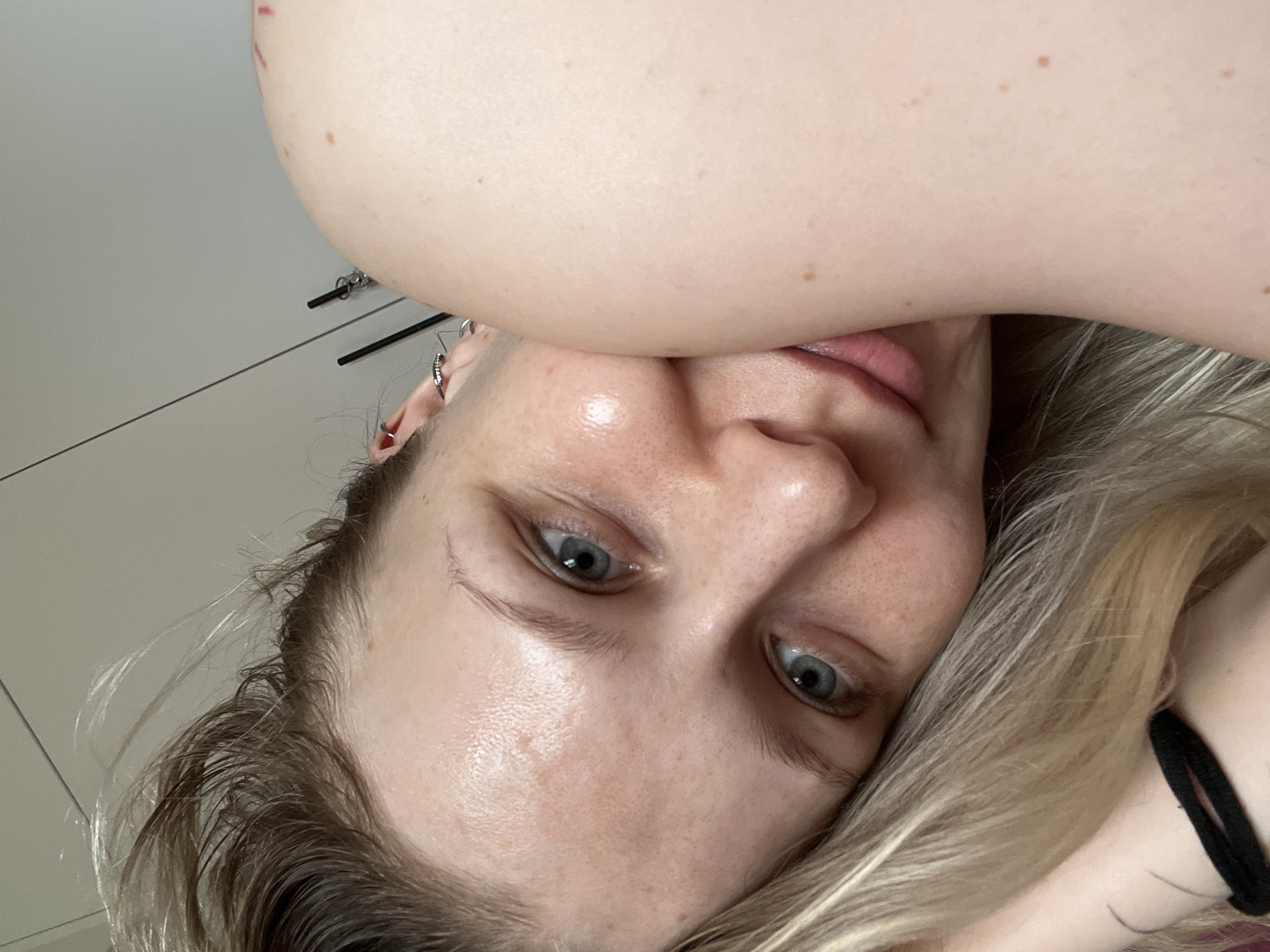 A close-up, upside-down shot of a fair-skinned person with blonde and brown hair. The person has grey eyes and is wearing earrings in their left ear. A smooth, pale arm is positioned above the person's forehead, partially obscuring the view. The background appears to be a plain white surface, possibly a wall. There is no evidence to indicate the location as Khimki, Russia.