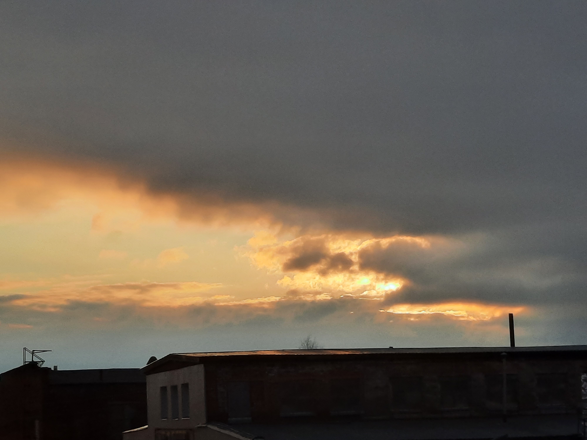 The image shows a cloudy sunset over an urban landscape. A portion of buildings is visible in the foreground. The sky is predominantly dark gray clouds, but the setting sun is visible in the distance. One building has three windows. The location is Krasnogorskoye, Russia.