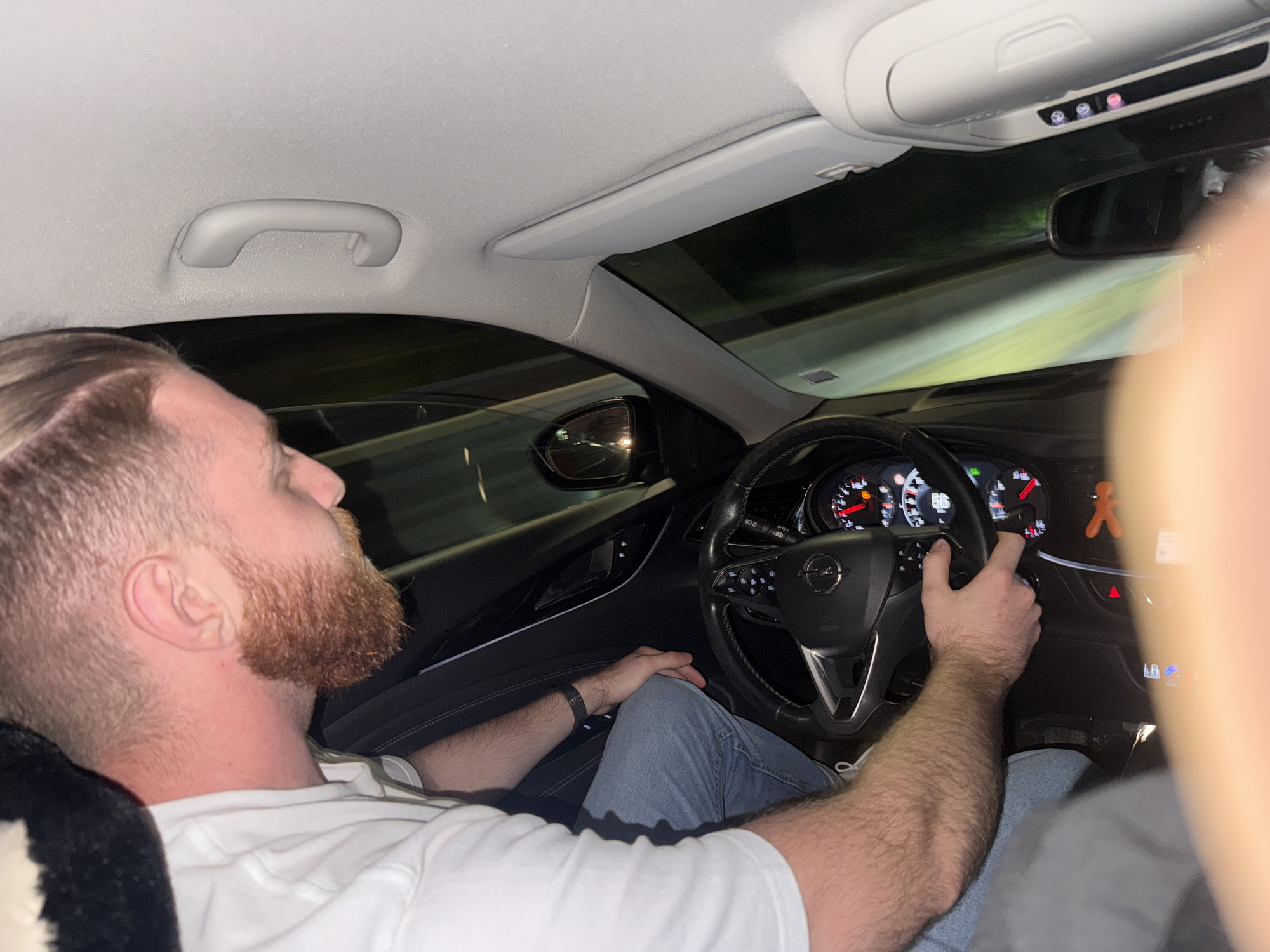 The image depicts the interior of a moving vehicle at night in the city of Varna, Bulgaria. A male driver, characterized by fair skin, an auburn beard, and short hair styled with faded sides, is seated behind the steering wheel. He is wearing a white t-shirt and light blue jeans, with a black wristband on his left arm. His hands are positioned on a black leather-wrapped steering wheel, which features an Opel logo in the center and integrated controls. The instrument cluster displays analog gauges, a digital reading of "86" (likely speed), and multiple illuminated warning lights.
The perspective suggests the photograph was taken by a front-seat passenger, whose blurred arm is visible on the right side of the frame. The car's interior includes a light gray headliner with a grab handle on the left and an overhead console with several buttons, two of which are illuminated in red/purple. Through the windshield and side mirrors, the exterior environment is dark, with streaks of green and yellow light indicative of vehicle motion on a road at night. A small orange, gingerbread man-like figure is hanging from the dashboard on the passenger side.