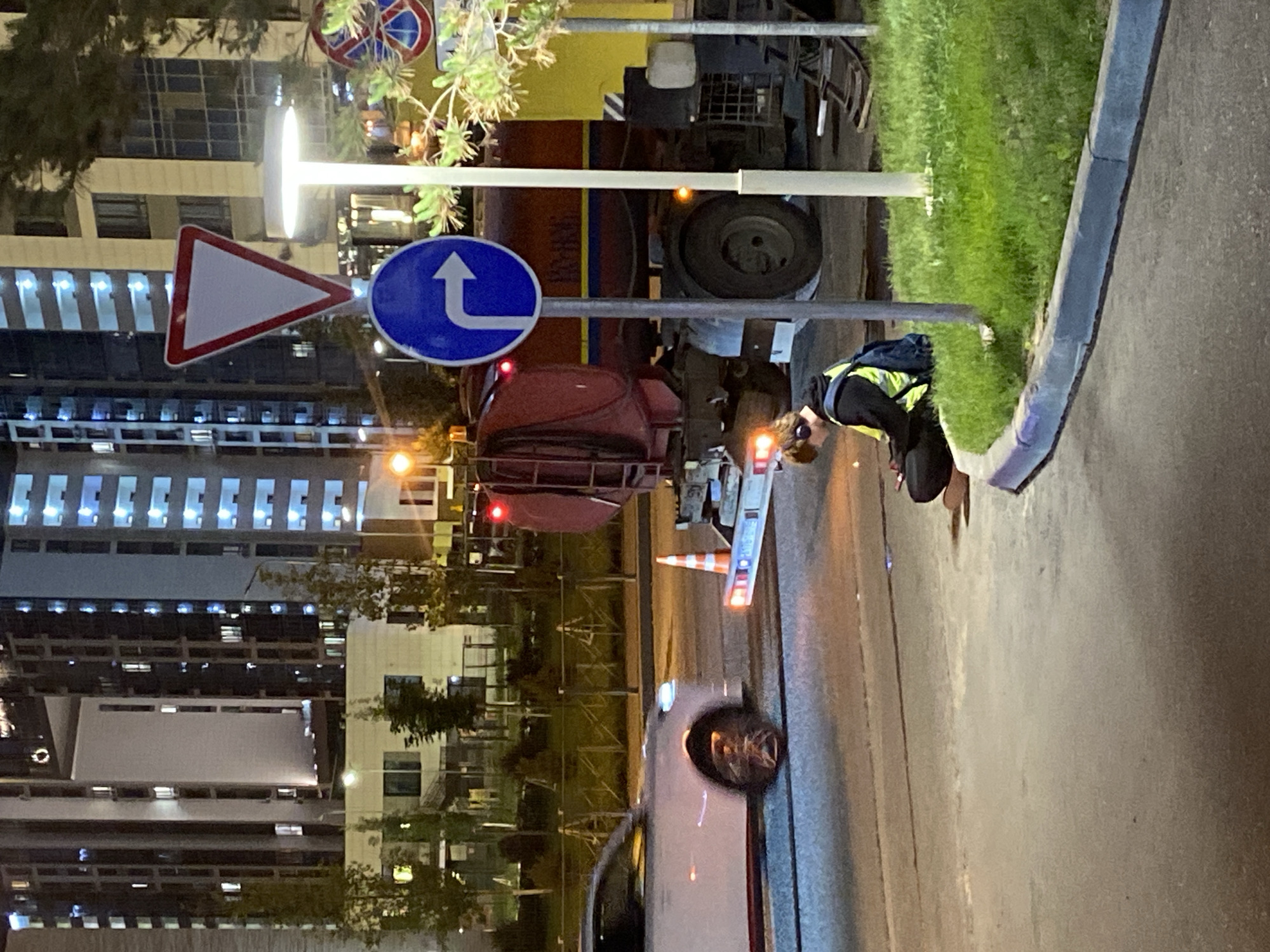 The image depicts a nighttime street scene in Kazan, Russia, characterized by artificial illumination. In the foreground right, an individual is seated on a concrete curb, wearing a dark hoodie, a high-visibility yellow vest, and a blue backpack. Over-ear headphones are visible, and a bright light emanates from their head area. They appear focused on an object in their hands.
To the left, a white sedan is partially visible, exhibiting motion blur on its front wheel and headlights, suggesting movement from right to left across the frame. In the mid-ground right, a stationary dark red utility truck, possibly a service vehicle, faces away from the viewer. It features yellow and blue stripes on its side, and its rear hazard lights are active, flashing orange/red. An orange traffic cone is positioned near its rear bumper.
Several traffic signs are prominent: a red and white inverted triangle "Yield" sign, a blue circular sign with a white right-turn arrow, and a partially obscured blue circular sign with a red diagonal line in the background right. A modern, disc-shaped LED streetlight stands upright on the right side of the road, illuminating a patch of green grass. Further back, multi-story buildings with numerous illuminated windows define the urban background, with one building featuring a distinctive large rectangular opening. A metal fence lines the left side of the road, behind the white car. The overall scene suggests an urban setting, possibly during or after a minor incident or service operation.