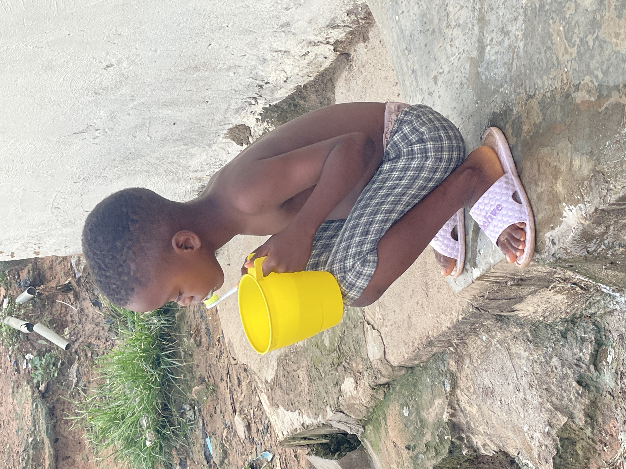 In the city of Abuja, Nigeria, a young child is crouched outdoors on a concrete ledge, holding a bright yellow mug in their left hand. The child is brushing their teeth with a white and green toothbrush, visible in their mouth. They are wearing plaid shorts and light purple slides. The background features a rough, textured wall to the right, and to the left, a dirt area with green grass and some scattered debris, including pieces of white pipe. The ground beneath the child appears wet and stained.
