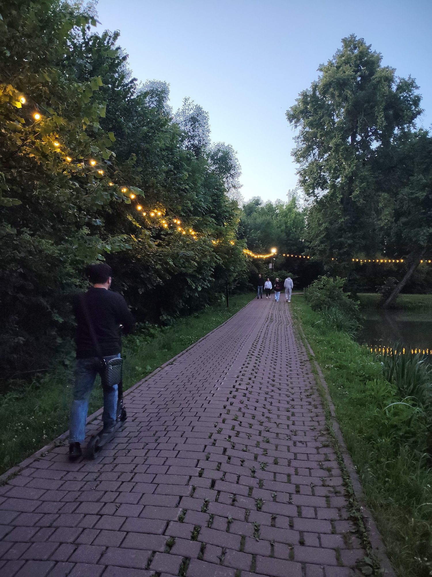 A man on a scooter travels along a paved path in a park, heading away from the viewer. He is wearing a black shirt, blue jeans, and a dark bag is slung across his back. The path is lined with trees and decorative string lights are visible along the path and in the trees. A pond or body of water is to the right of the path. In the distance, several people are walking along the path. The sky is a soft, pale blue, indicating dusk or dawn. This scene is set in the city of Chekhov, Russia.