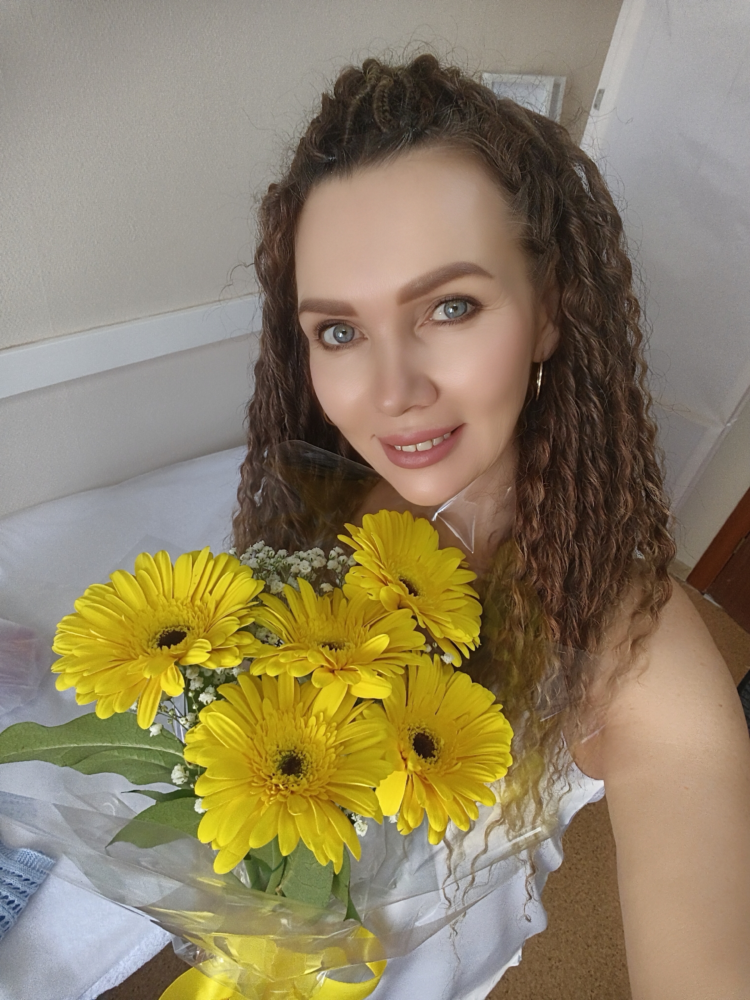 A woman is holding a bouquet of yellow gerbera daisies in front of her. She has light skin, blue eyes, and light brown curly hair with some braids on the top of her head. She is wearing gold hoop earrings and smiling. The background appears to be a room with a bed. Location: Unknown, Unknown.