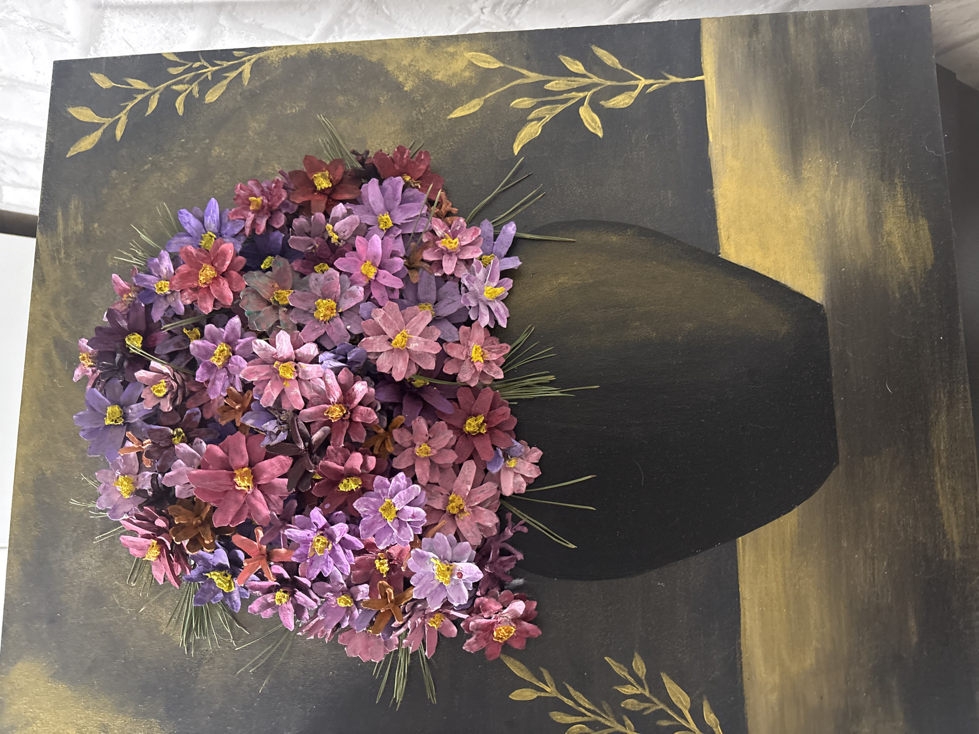 The image displays a still life artwork featuring a vase of flowers on a painted surface. The vase is black and appears to be made of a smooth material. It holds a large, full arrangement of small, multi-colored flowers in shades of pink, purple, and reddish-brown, with bright yellow centers. Green pine needles are interspersed within the floral arrangement, adding a touch of natural texture.
The background of the artwork is dark with a textured, mottled effect, predominantly in shades of black and dark grey, with hints of gold. There are decorative gold leaf sprigs or branches painted on either side of the vase, adding an elegant touch to the composition. The vase sits on a horizontal surface that is painted with gold and dark tones, suggesting a table or shelf. The overall style of the artwork is decorative and somewhat rustic, likely created using mixed media or natural elements. There are no people present in the image, and the artwork itself is the sole subject. The location of this artwork within the city of Omsk, Russia, cannot be determined from the visual information provided in the image.