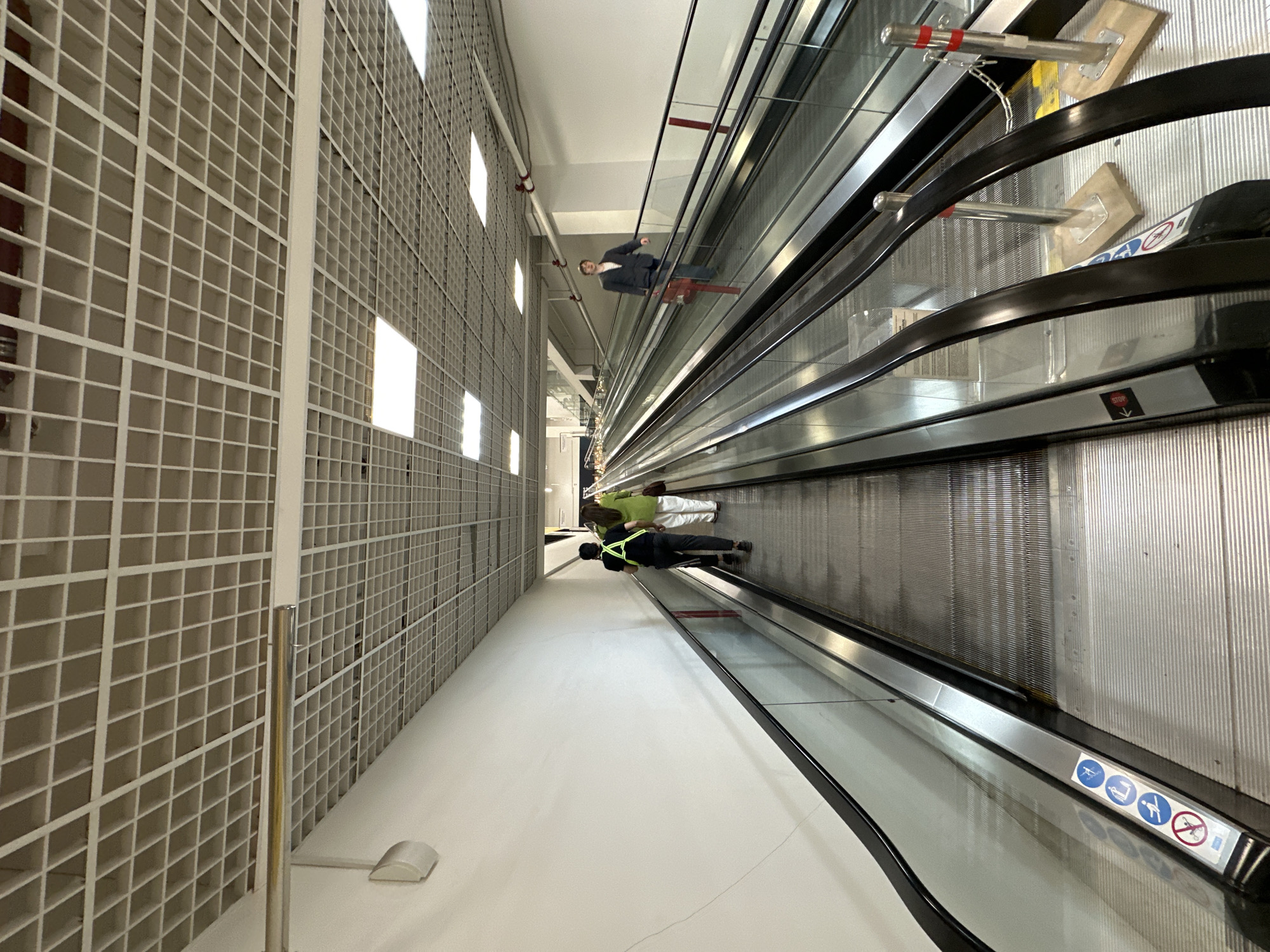 The image depicts an indoor scene, likely a commercial or transportation hub in Moscow, Russia. The primary focus is on several moving walkways (travelators) stretching into the distance. The travelators are modern, with black handrails and prominent safety signage at the entrance indicating prohibitions such as using strollers, carrying luggage, or running.
Several individuals are present on the travelators. One person, possibly a businessman, is moving upward, while two people, possibly maintenance workers, are seen further down.
A distinctive feature of the environment is the industrial-style wall on the left, consisting of a white grid pattern. The floor is bright white, contributing to a clean and modern aesthetic. The lighting is artificial and bright.