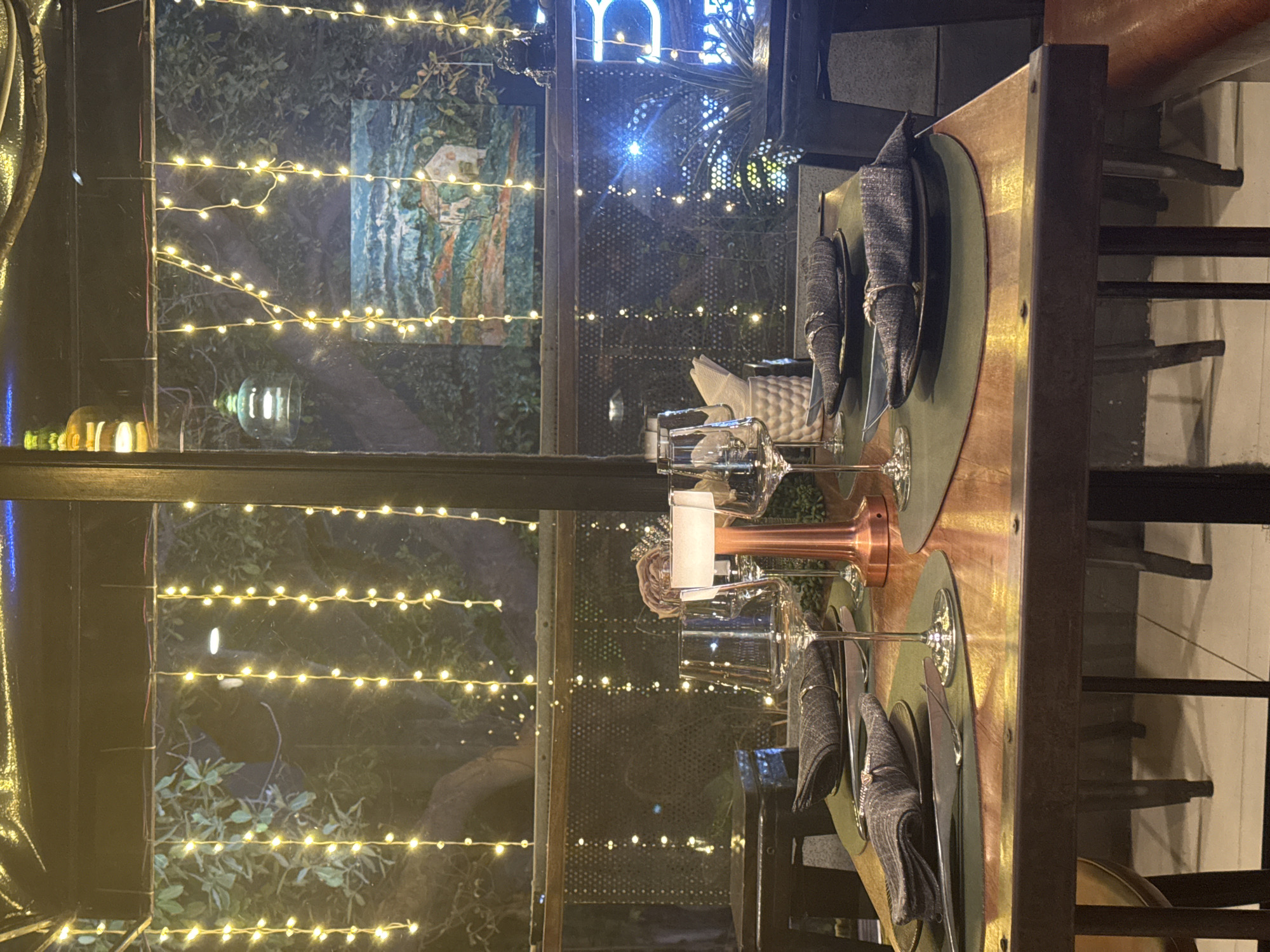 The image shows a dining table set for a meal in a dimly lit restaurant. The table is made of wood with a metal frame. On the table, there are green placemats, plates with folded dark gray napkins secured with rings, and cutlery. Several wine glasses and a copper-colored lamp are also on the table. In the background, there is a large window looking out onto a garden with trees. String lights are draped across the window, creating a festive atmosphere. There is a painting on the wall behind the window, depicting a landscape with a house. The restaurant appears to be decorated in a modern style with a touch of elegance. The overall impression is of a cozy and inviting dining experience. It is not possible to determine the specific location in Hanoi, Vietnam from the image.