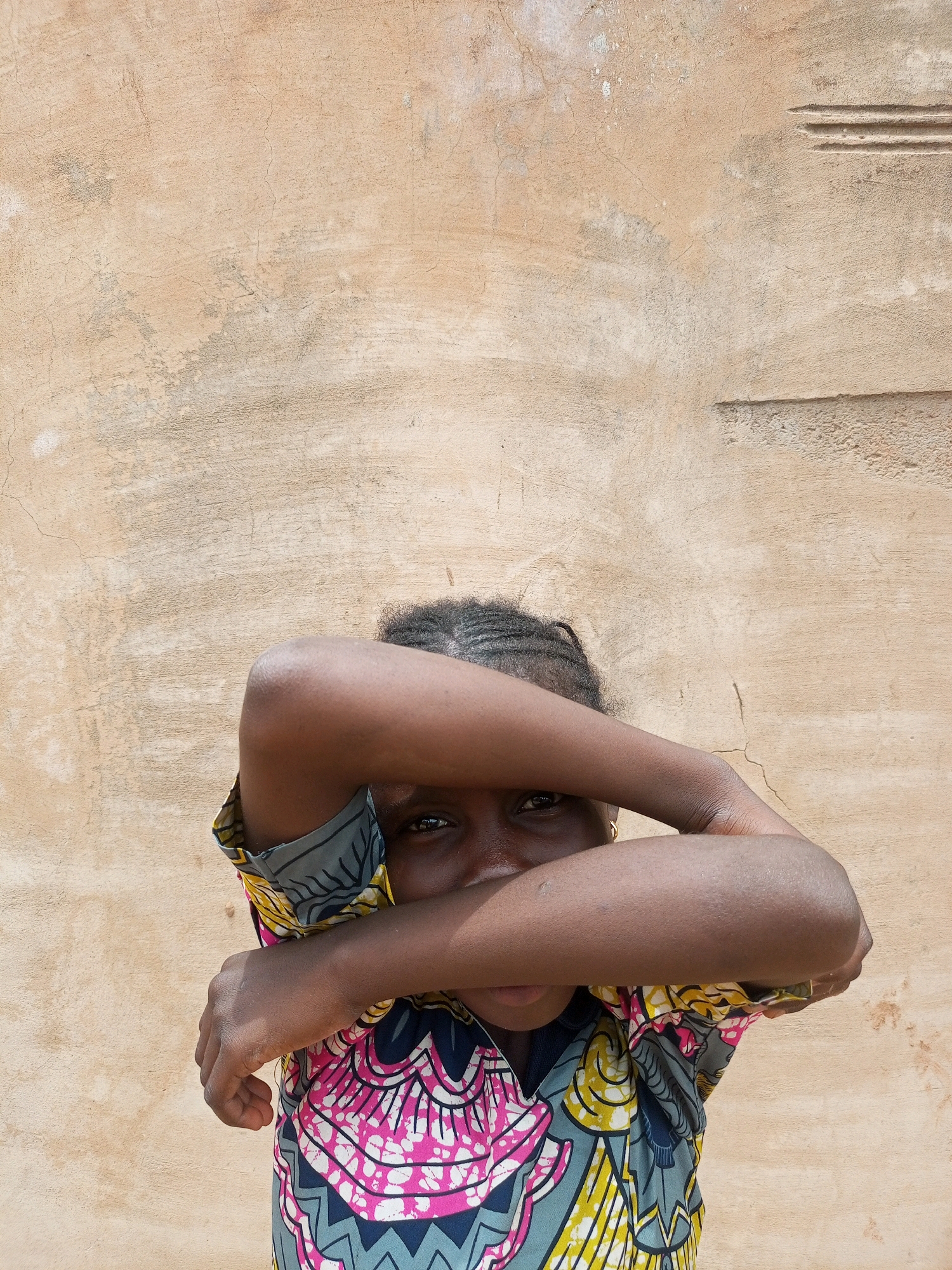 A young girl with braided hair stands in front of a textured, light brown wall in Gombe, Nigeria. She is wearing a vibrantly patterned shirt with pink, yellow, grey, and blue designs. Her arms are crossed in front of her face, obscuring most of her features except for one eye. The wall behind her has some cracks and inconsistencies in its surface.