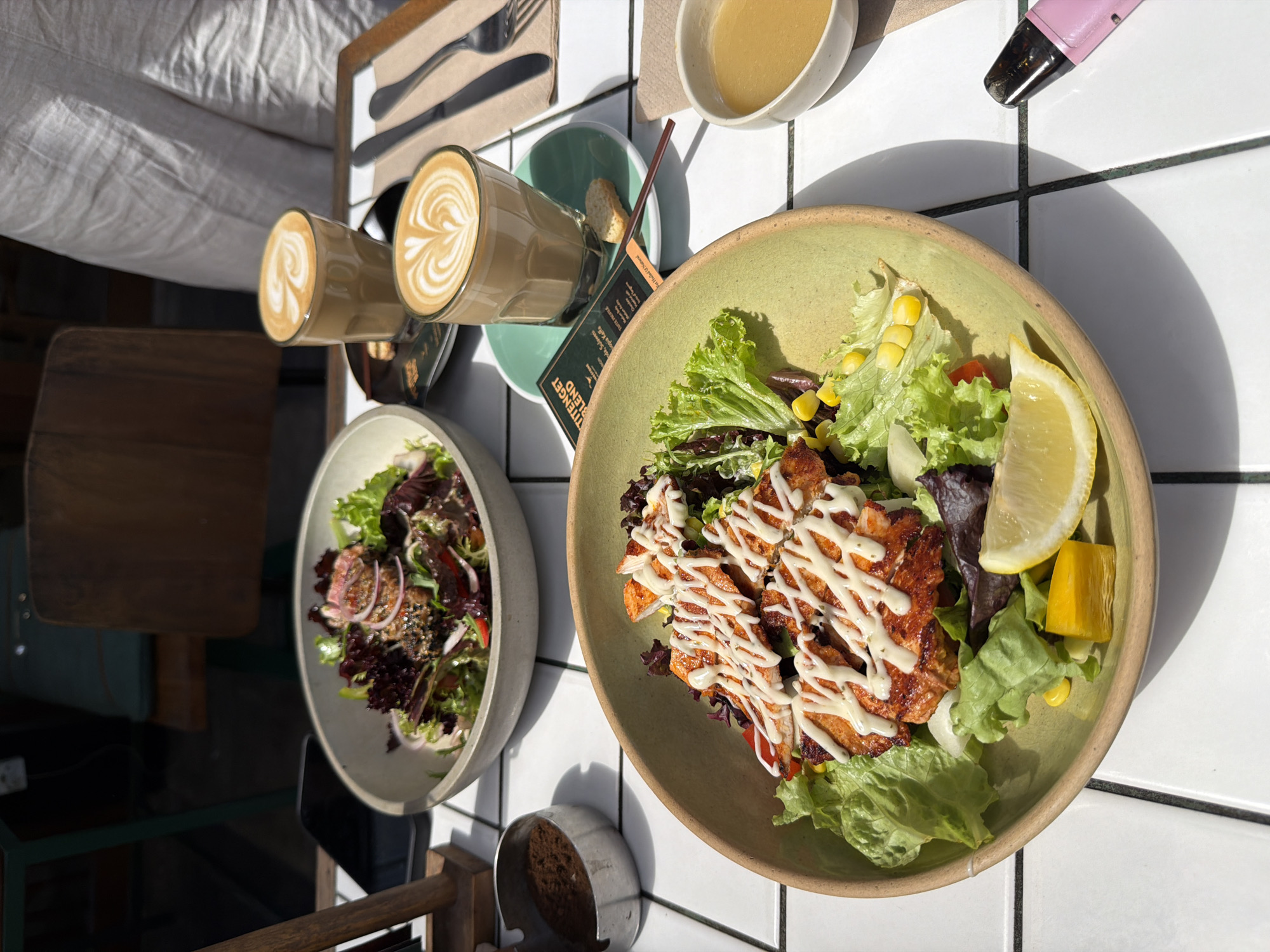 On a white tiled table, two plates of salad and two cups of coffee are arranged. The salad in the foreground is in a light green bowl and contains grilled chicken, lettuce, corn, a slice of lemon, and a piece of diced yellow vegetable. The second salad, in a gray bowl in the background, appears to have slices of meat, red onion, and various greens. Two cups of coffee with latte art sit on saucers. One coffee cup is on a dark blue saucer, and the other is on a green saucer. Next to the coffees, a small white cookie rests on a napkin. To the right of the coffee cups, a fork and knife are placed on a napkin. In the upper left, a metal dish contains a dark substance, possibly coffee grounds. A pink electronic device with a black tip is on the table near the foreground salad. The scene is set outdoors, with a glimpse of chairs and a table in the background, suggesting a cafe or restaurant in North Kuta, Indonesia. The sunlight casts shadows on the table.
