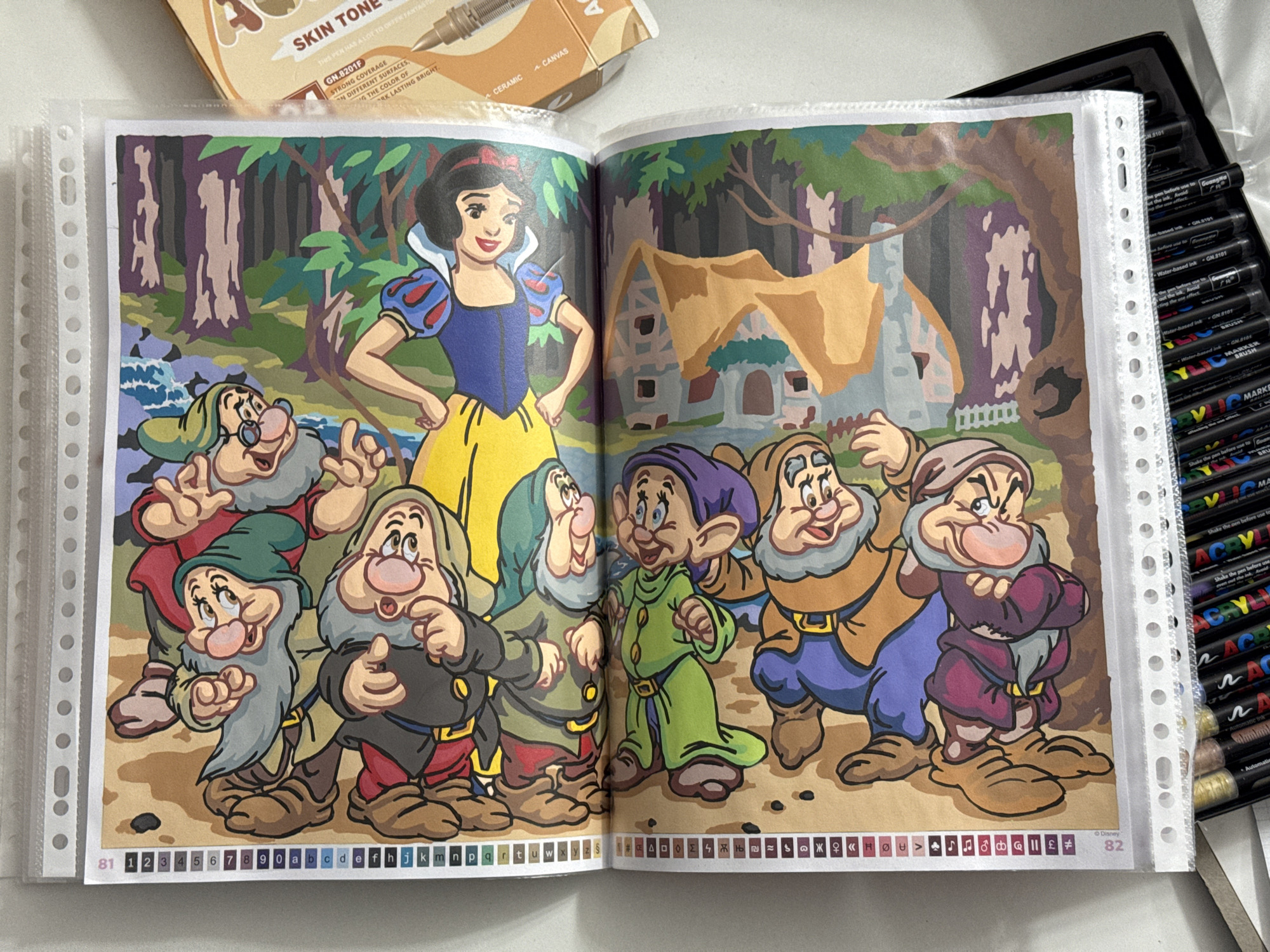 The image shows two pages of a coloring book featuring characters from Disney's Snow White and the Seven Dwarfs. The left side of each page has a color-coding guide with numbers and letters. The top page depicts Snow White with six of the dwarfs in a forest setting, while the bottom page shows a different arrangement of the dwarfs, seemingly in front of buildings. The pages are housed in a plastic binder. To the right of the coloring book pages, part of a box and a marker pen are visible. The marker pen is labeled "SKIN TONE". The image is taken indoors, with a white wall visible in the background. There is no visible information to suggest the location is in Łódź, Poland.