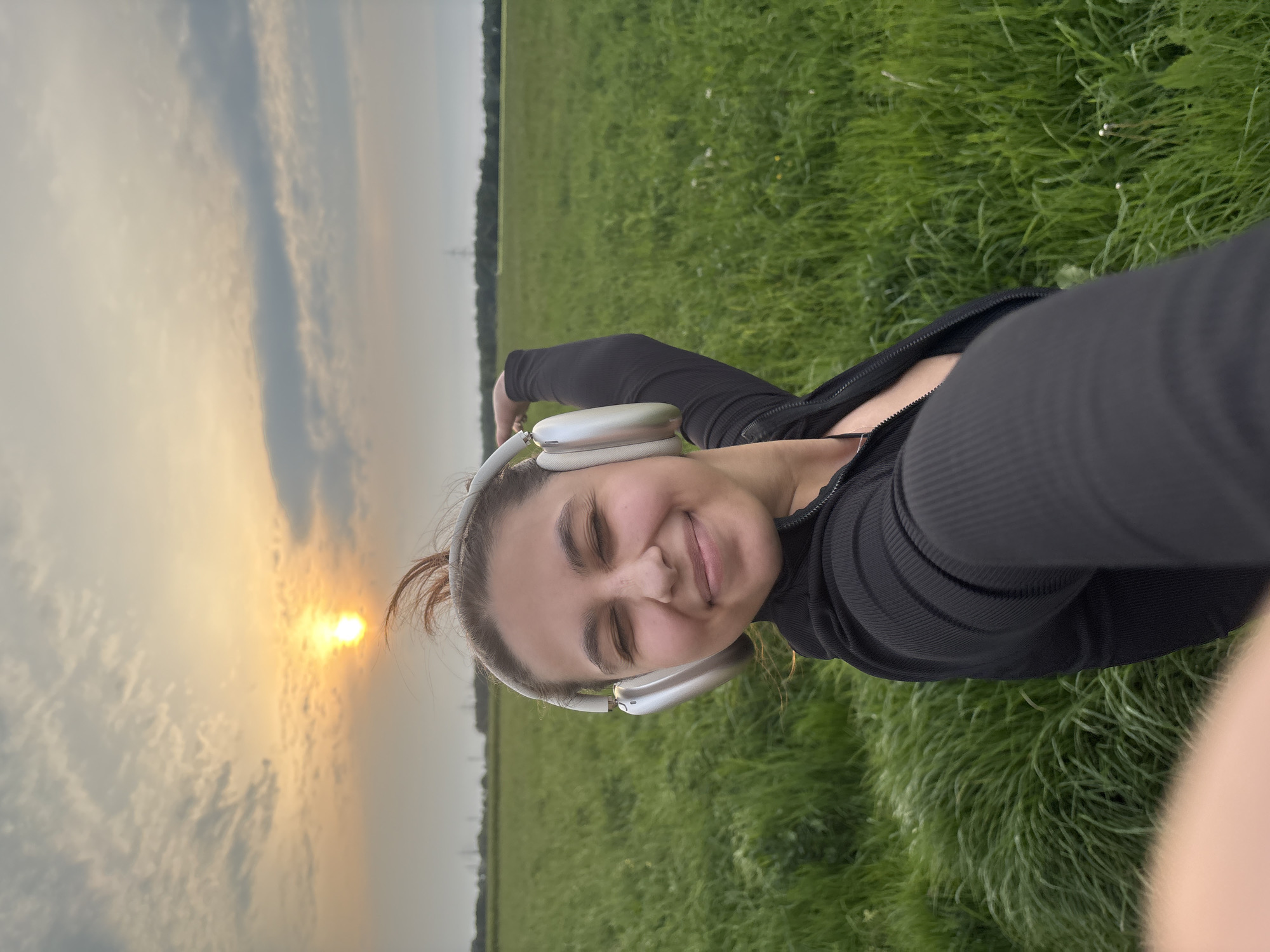 A young woman with fair skin is wearing headphones and a black zip-up shirt and is lying in a field of green grass. She is smiling with her eyes closed and appears relaxed. The sky is visible in the background with a setting sun. It's dusk. Given the landscape, it's likely the scene is near a rural area of Mönchengladbach, Germany, though there's no specific landmark visible to confirm the exact location.
