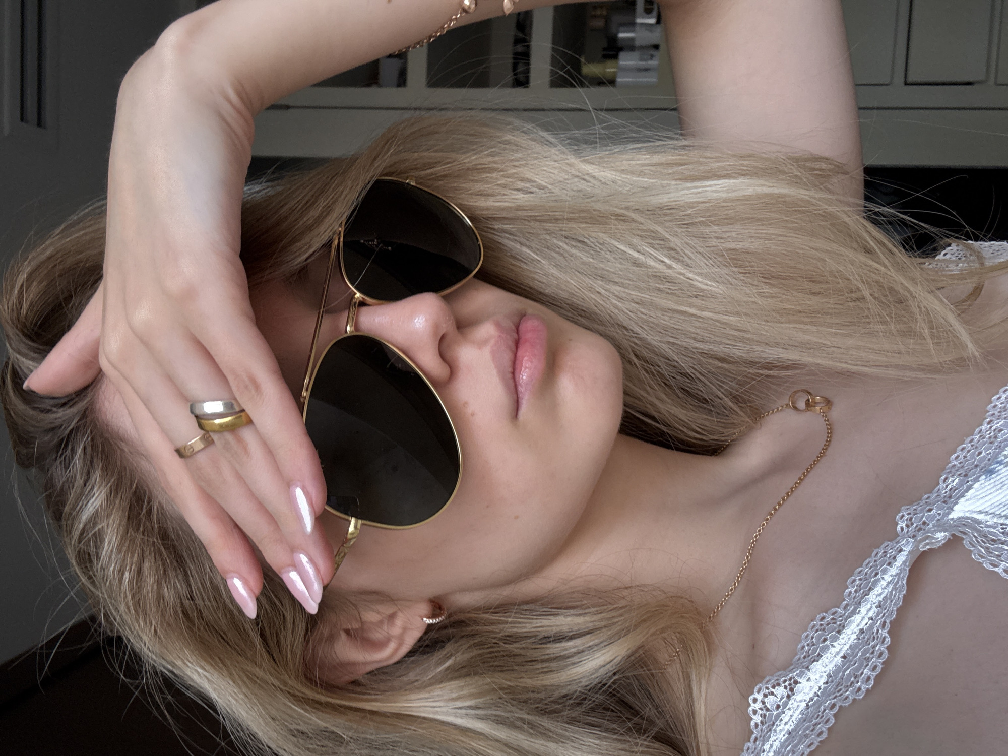 This close-up portrait, taken in Mexico City, Mexico, features a person, likely a woman, with long, light blonde hair that is spread out around her head. She is seen from a slightly elevated angle, with her head tilted to the side, appearing to be lying down. She wears large, gold-framed aviator sunglasses with dark lenses that obscure her eyes.
Her left arm is raised, with her hand resting gently near her head and covering part of her forehead. Her nails are long, almond-shaped, and painted with a glossy, light pink, pearlescent polish. On her left hand, she wears several rings: one finger has both a wider silver-toned band and a thinner gold-toned band, while another finger has a slender gold-toned band. A delicate gold chain necklace with a small circular accent hangs around her neck, and a small hoop earring with visible sparkle is in her right ear. Her lips are slightly parted, revealing a soft, natural pink color.
A white garment with intricate lace detailing is visible on her right shoulder and chest, suggesting a stylish top or lingerie. The background is dark and out of focus, providing no specific location cues beyond the implied indoor or controlled setting. The overall mood conveyed is one of relaxed elegance and fashion. No text is visible in the image.