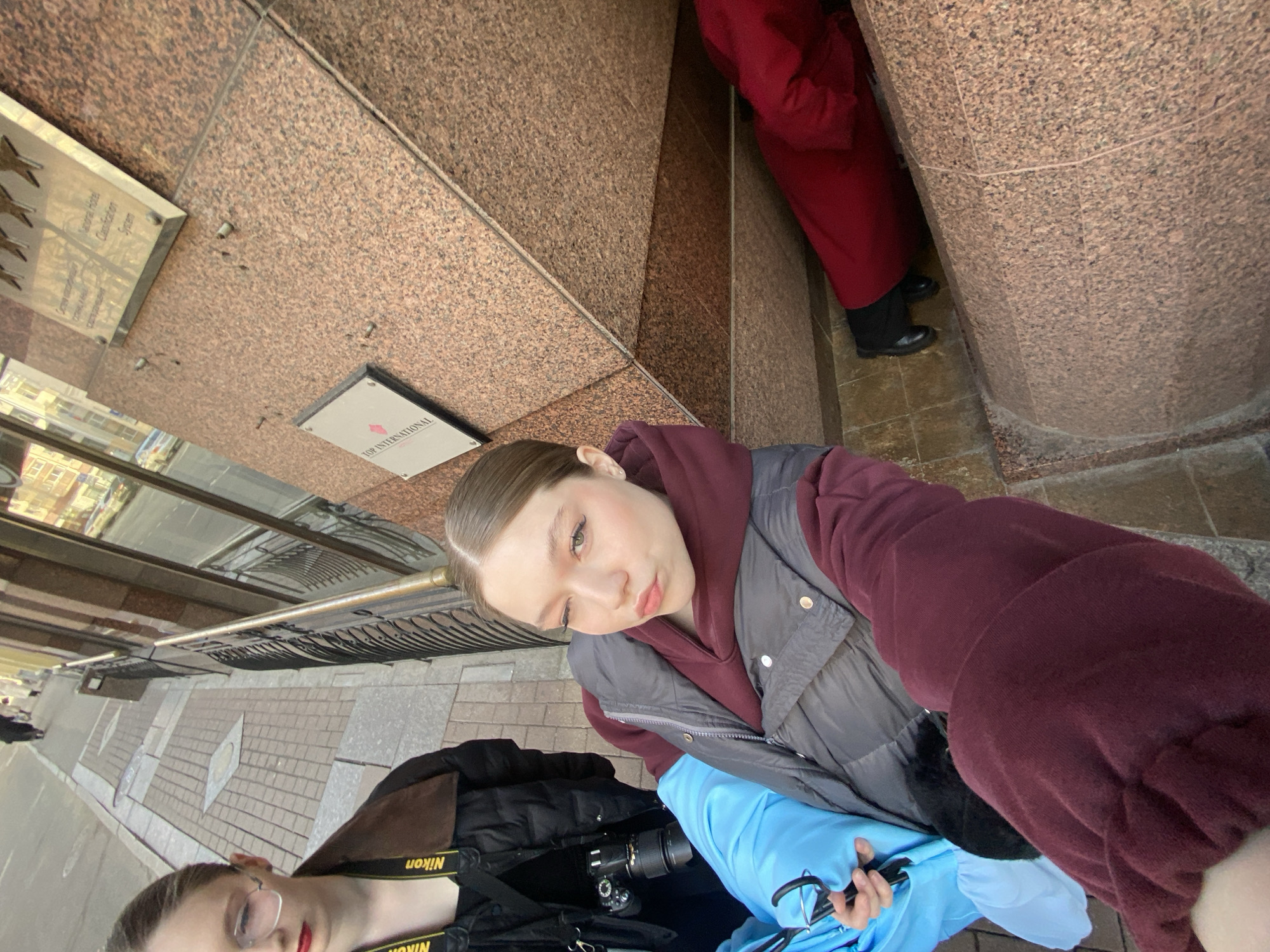 This image depicts a street-level scene in the city of Saint Petersburg, Russia, focusing on three individuals in front of a building identified by prominent signage. The building's facade is constructed from polished reddish-brown granite-like stone tiles. Affixed to the facade are multiple signs: a larger metal plaque displaying five gold stars and text in both English ("National Hotel Classification System") and Cyrillic, and a smaller white rectangular sign clearly stating "THE INTERCONTINENTAL" with a red diamond logo. A partially visible third sign reads "TOP INTERNATIONAL."
The central figure, a young female with light brown hair pulled back, occupies the foreground, extending her right arm to capture a selfie. She is dressed in a maroon hooded sweatshirt under a dark grey puffer vest. Her facial expression includes a wink of her right eye and pouted lips. Her left hand is holding a light blue garment and a black wire hanger.
To the left of the central figure, a second young female is partially visible from the chest up. She wears clear-framed glasses, red lipstick, a black puffer jacket over a black top, and a brown collar. A yellow "Nikon" branded camera strap with an attached black DSLR camera and lens hangs around her neck. Her gaze is directed downwards and to the left, away from the primary camera.
In the lower right quadrant of the image, the lower legs and black-shod feet of a third individual, clad in red trousers or a long skirt, are visible, positioned low to the ground against the building's base.
The sidewalk is paved with grey and darker grey interlocking bricks arranged in a geometric pattern. In the background, a street scene is discernible, featuring reflections of other buildings in the glass windows of the structure and showing pedestrians on the street beyond. The scene appears to be illuminated by bright, ambient daylight.