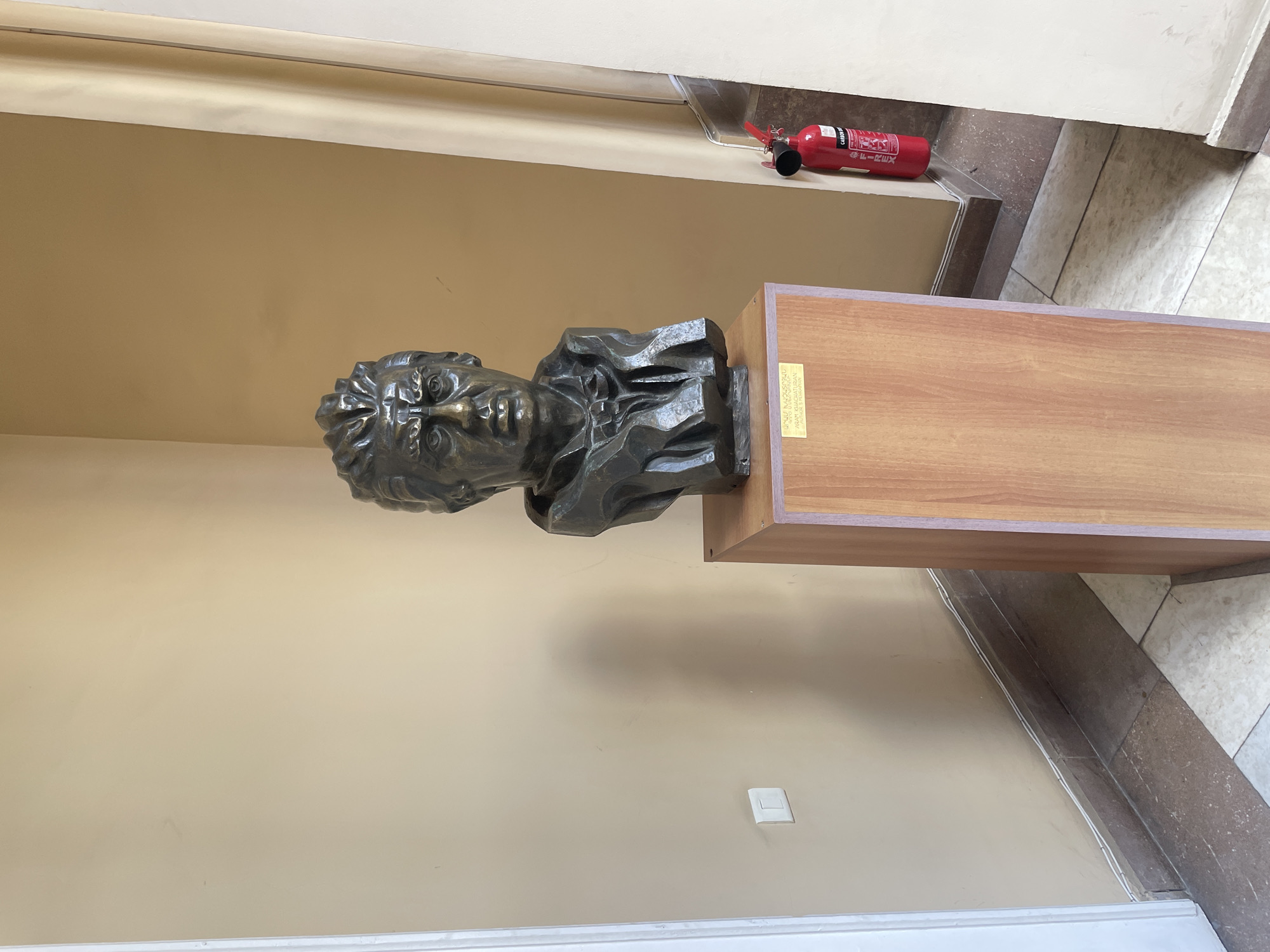 The image shows a bust of Komitas Vardapet, an Armenian priest, musicologist, composer, arranger, singer, and music pedagogue, in a public setting. The bust is made of dark metal and is mounted on a wooden pedestal. There is a plaque on the front of the pedestal that contains information about Komitas Vardapet. The image was taken in Yerevan, Armenia. A fire extinguisher is visible on the wall above the bust. The floor is tiled. The wall is painted a light beige color. A small electrical outlet is visible on the wall below the bust.