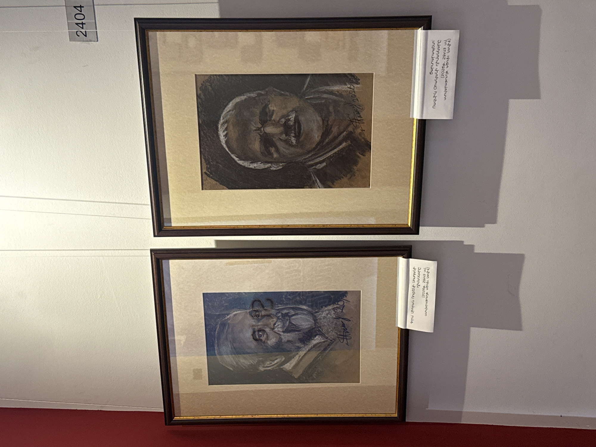 Two framed portraits hang on a light-colored wall, situated above a red surface that appears to be a floor or base. The top portrait features a subject with a dark, smudged style, possibly charcoal or a similar medium. The lower portrait depicts a face in predominantly blue tones. Each portrait is framed in dark wood with a light-colored matte. To the left of the top portrait is a sign with the number "2404". To the right of each portrait, a small white paper appears to be attached. The writing on the papers is illegible. The location is in Yerevan, Armenia.