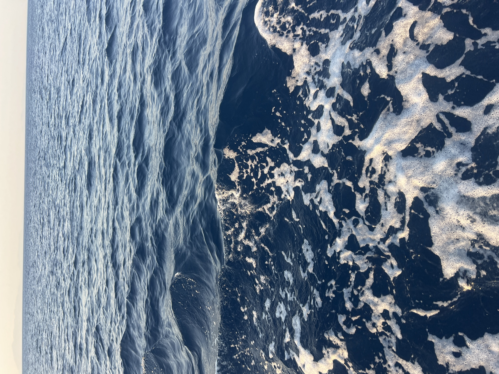 The image depicts a close-up view of the ocean's surface, with a distinct line separating calm water from choppy, foamy water. The calm water, in the upper portion of the image, is a deep blue with gentle ripples reflecting the light. The choppy water, in the lower portion, is a darker blue and is filled with white foam and bubbles, indicating movement and disturbance, likely from a boat. The horizon is visible in the distance, a faint line separating the sea from a pale sky. There are no people or man-made objects clearly visible in the immediate scene. The overall impression is one of motion and the natural beauty of the ocean. The location is Unknown, Unknown.