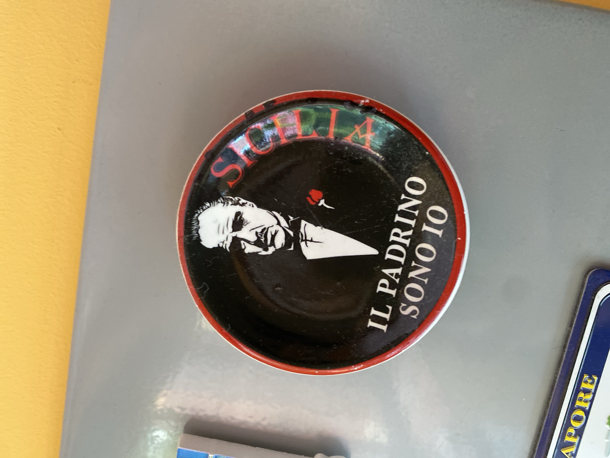 The image shows a close-up, eye-level shot of a magnet on a refrigerator door. The magnet is oval-shaped with a red and black design. It features a black and white illustration of a man in a suit and bow tie, with a red rose on his lapel. The text on the magnet reads "IL PADRINO SONO IO" and "SICILIA". The magnet is situated on a grey refrigerator door, which is partially illuminated by the yellow wall behind it. The overall setting suggests a domestic environment. The provided information does not include the city of Wrocław, Poland.