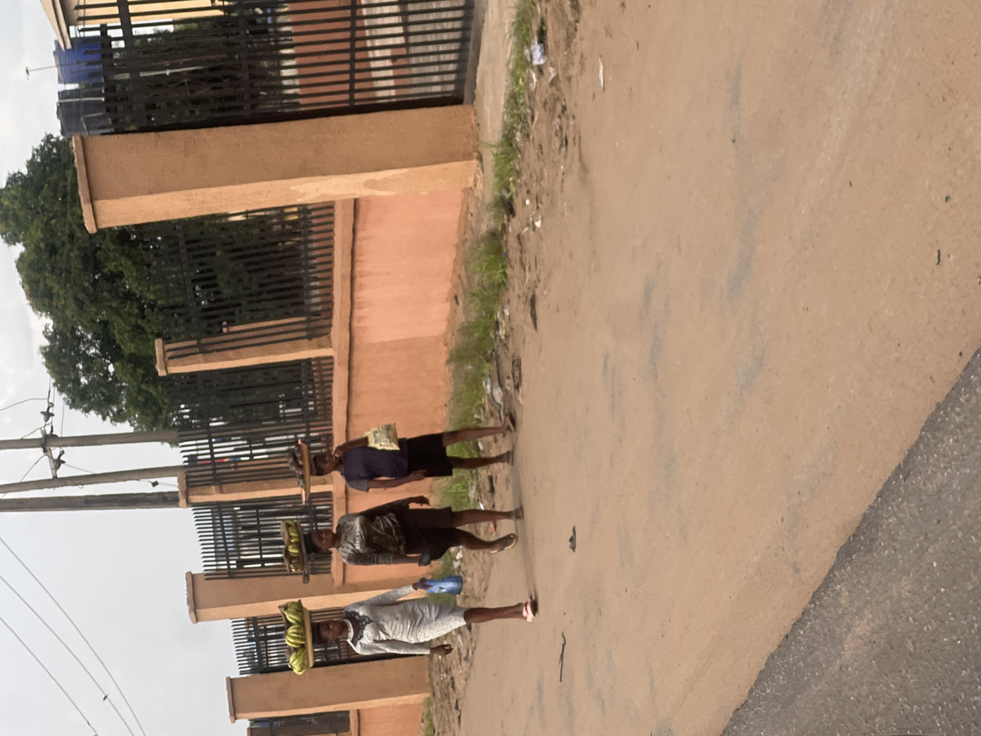 Three people are walking along a dirt road in Unknown, Unknown, each carrying items on their head. The backdrop consists of a fence, a building and a tree. The sky is overcast.
