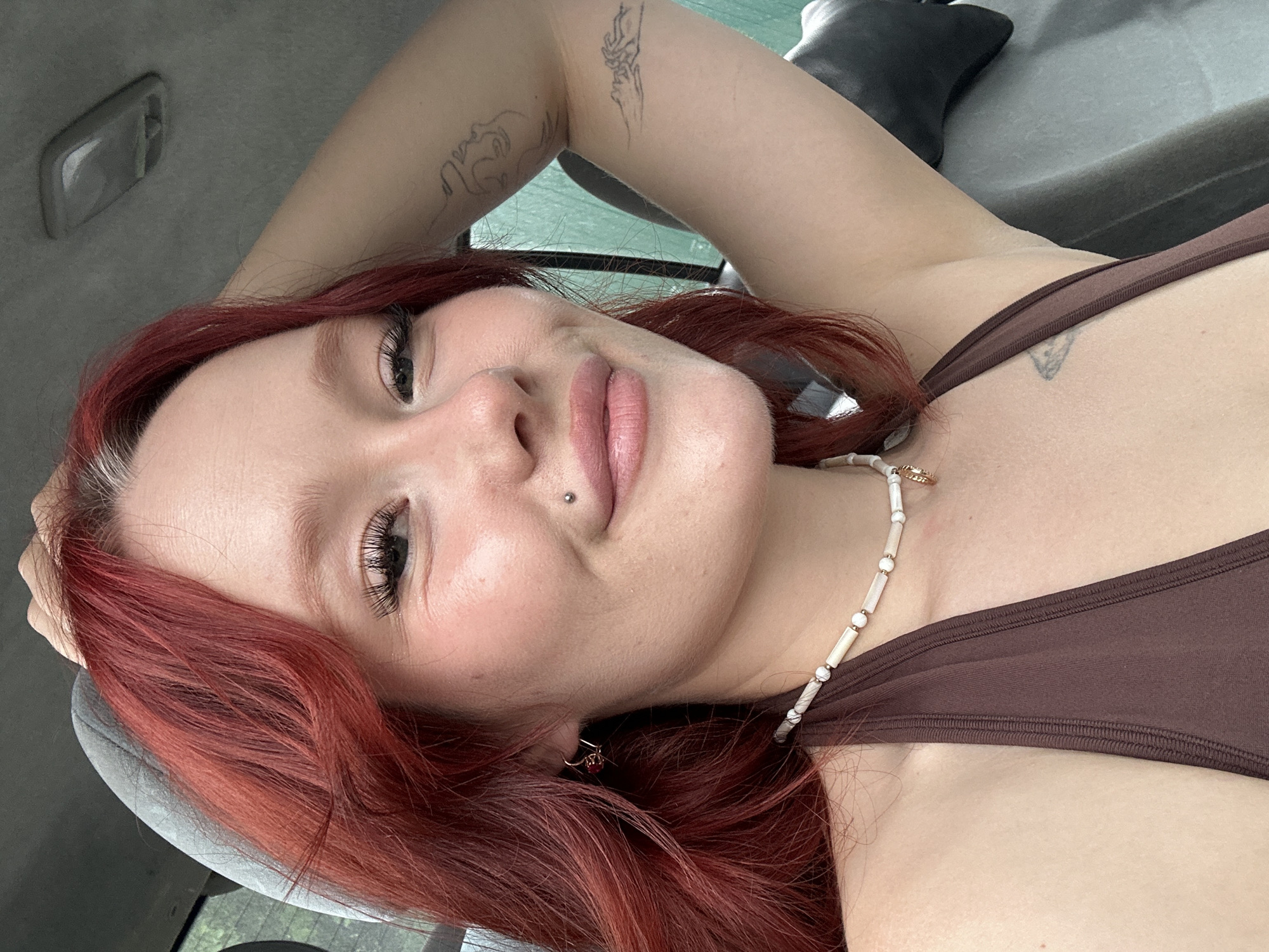 The media file is a close-up, rotated selfie of a person with red hair. The subject is lying down, with their head tilted back and one arm resting behind their head. Their eyes are open, and they are looking directly at the camera with a relaxed expression. The person has full lips, accentuated by pink lipstick, and a lip piercing. They are wearing a brown tank top and a beaded necklace. There are small tattoos visible on their arm and chest. The background appears to be the interior of a car, with a gray headliner and a black headrest visible. The lighting suggests it is daytime. There is no visible text.