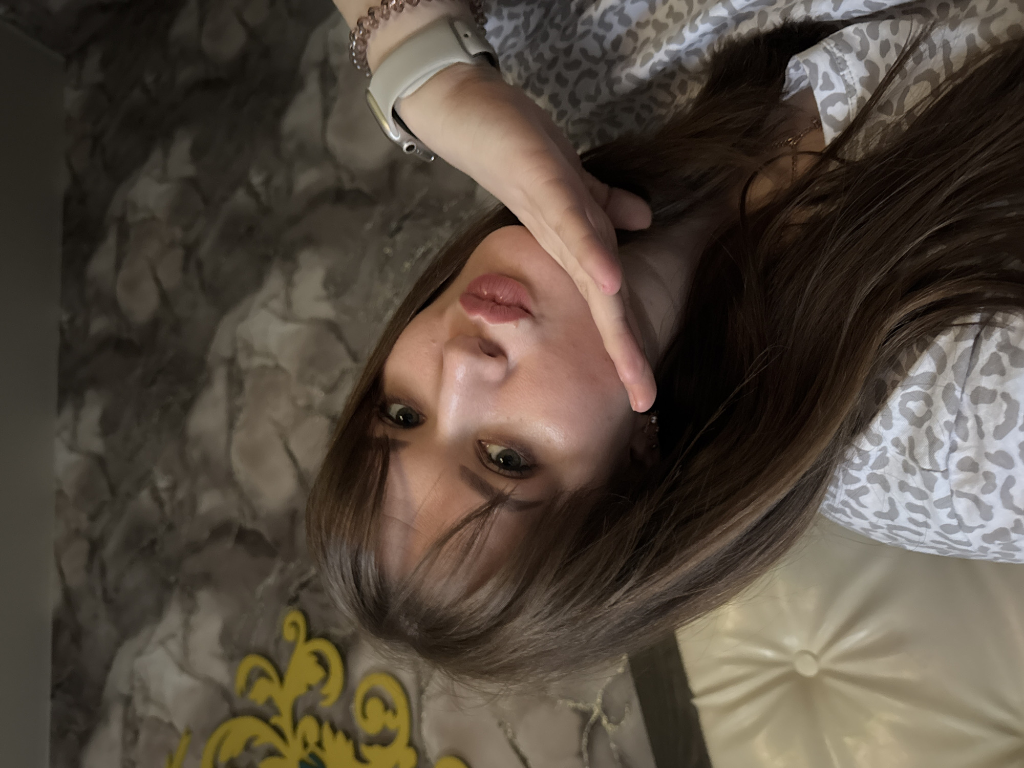 A woman with brown hair is pictured indoors. She is wearing a smartwatch with a white band on her left wrist. She is wearing a patterned top. The background includes a textured wall and a padded surface. The picture was possibly taken in Chaikovsky, Russia.