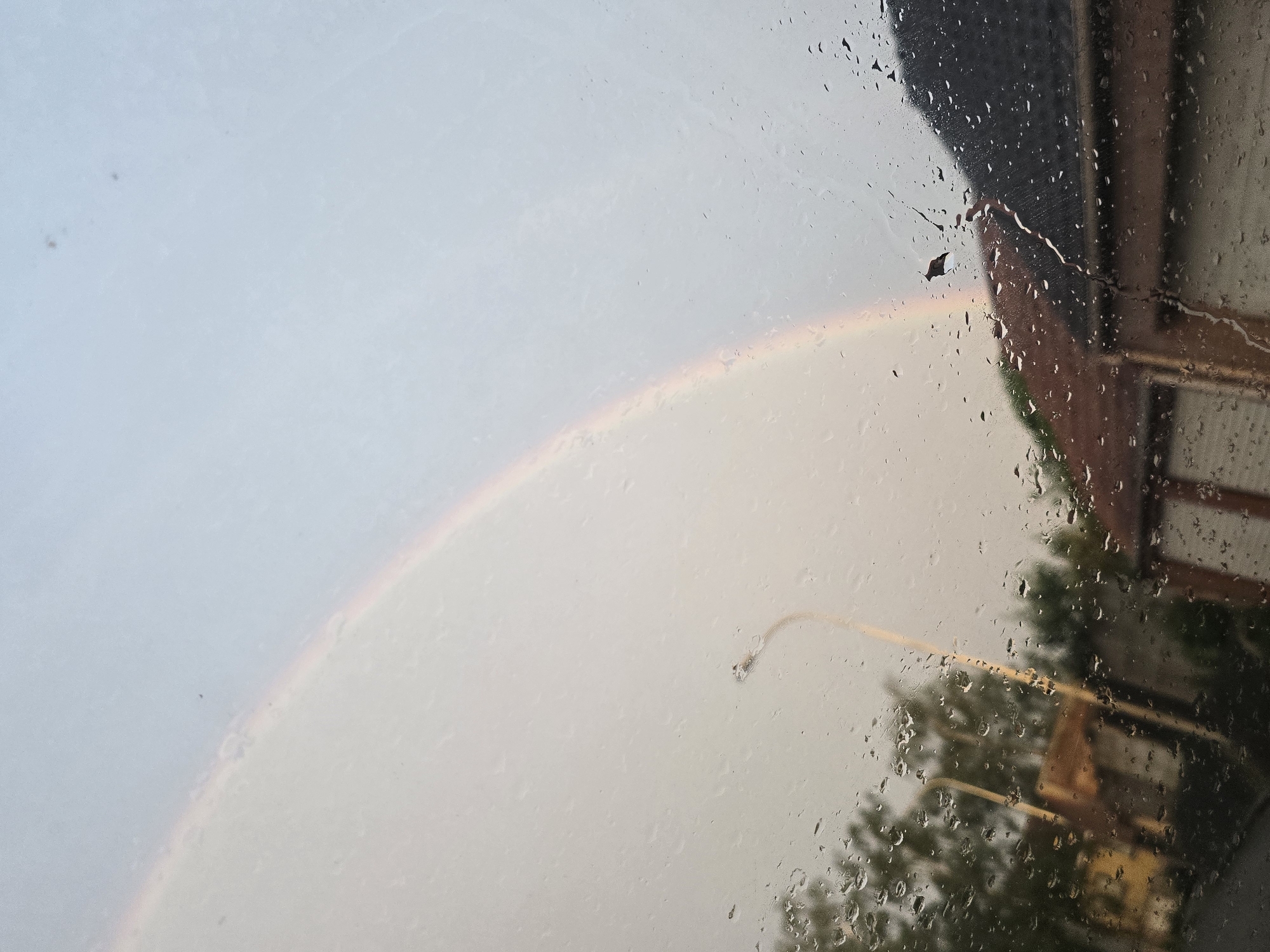 The image presents an outdoor scene viewed through a rain-streaked window, from an apparent indoor vantage point. Numerous water droplets of varying sizes cover the glass, creating a blurred and distorted perspective of the exterior.
The most striking feature is a partial rainbow, visible as a faint arc of color spanning from the middle to the lower-left portion of the frame. The sky beyond the window appears overcast and light, with hints of grey and pale blue, suggesting recent or ongoing rainfall coupled with sunlight breaking through.
Through the rain-covered glass, details of the surroundings are discernible. In the upper right quadrant, a section of a building is visible, characterized by a dark, possibly tiled or shingled roofline or overhang, and a lighter, textured facade that might be brick or wood. A small piece of debris, possibly a leaf, clings to the window in this area.
In the lower right, a distinct, curved, yellow-orange object, consistent with the arm of a street lamp, is apparent. Beneath and around this, blurred dark green shapes indicate the presence of trees or dense foliage.
The scene conveys the atmosphere of a rainy day, likely during the late afternoon or early evening given the formation of the rainbow. No people are visible, and there is no discernible text in the image. The specified location for this image is Mouscron, Belgium.