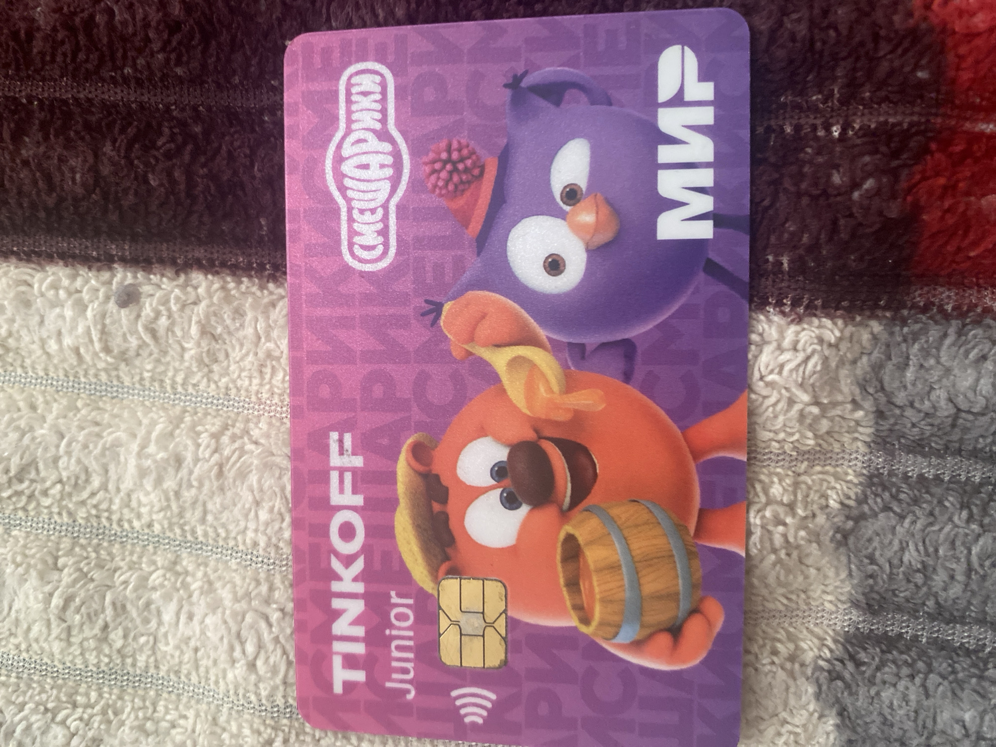 The image shows a Tinkoff Junior debit card with characters from the animated series "Smeshariki" (also known as "Kikoriki"). The card is pink and purple with these characters depicted. The Tinkoff logo is visible in white at the top left, and the word "Junior" is below it. The "Smeshariki" logo is in white on the right side of the card. The card also features a chip and contactless payment symbol. The card is placed on a textured surface, likely a blanket, with a pattern of cream and dark brown stripes. There is no information in the image to indicate its location in the city of Kizilyurt, Russia.