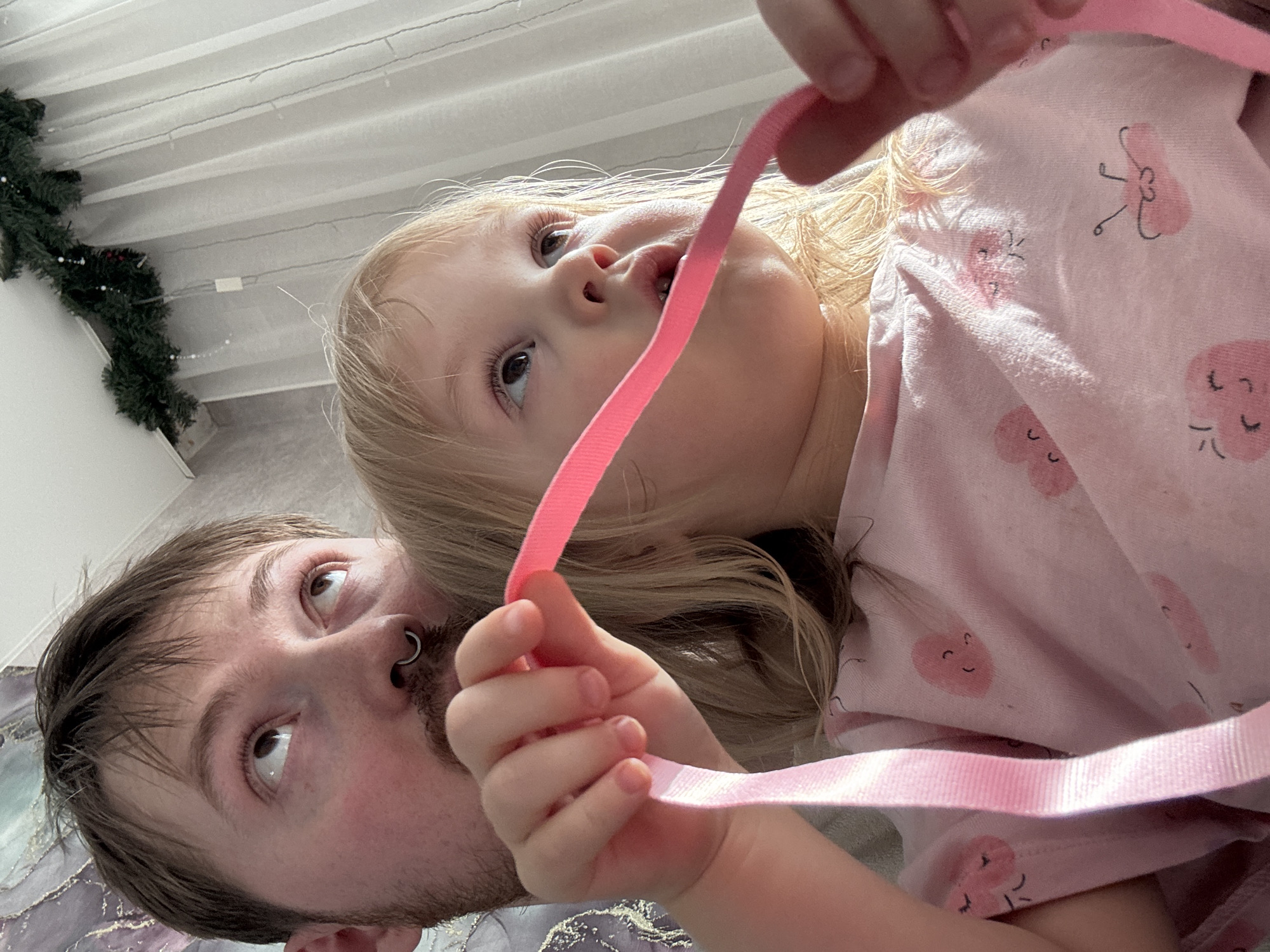 A low-angle, close-up indoor shot captures a man and a young child lying down and looking upwards. The man, positioned on the left, has brown hair, light stubble, and a silver septum piercing. His eyes are directed upwards and to his right.
To his right, a young child with blonde hair is prominent. She is wearing a light pink top patterned with cartoon-like heart shapes, some depicting small smiling faces. The child holds a bright pink ribbon in her hands, parts of which are near her mouth. Her gaze is also directed upwards and slightly to the left.
The background shows a white ceiling with linear details, strung with delicate string lights. A dark green decorative garland is visible in the upper left corner. A patterned fabric, likely a blanket or sheet, is beneath the man. The lighting is soft and even, suggesting a comfortable indoor environment. No text is visible in the image. Both subjects display neutral or slightly curious expressions as they look towards an unseen point above them. The scene is set in Chaikovsky, Russia.