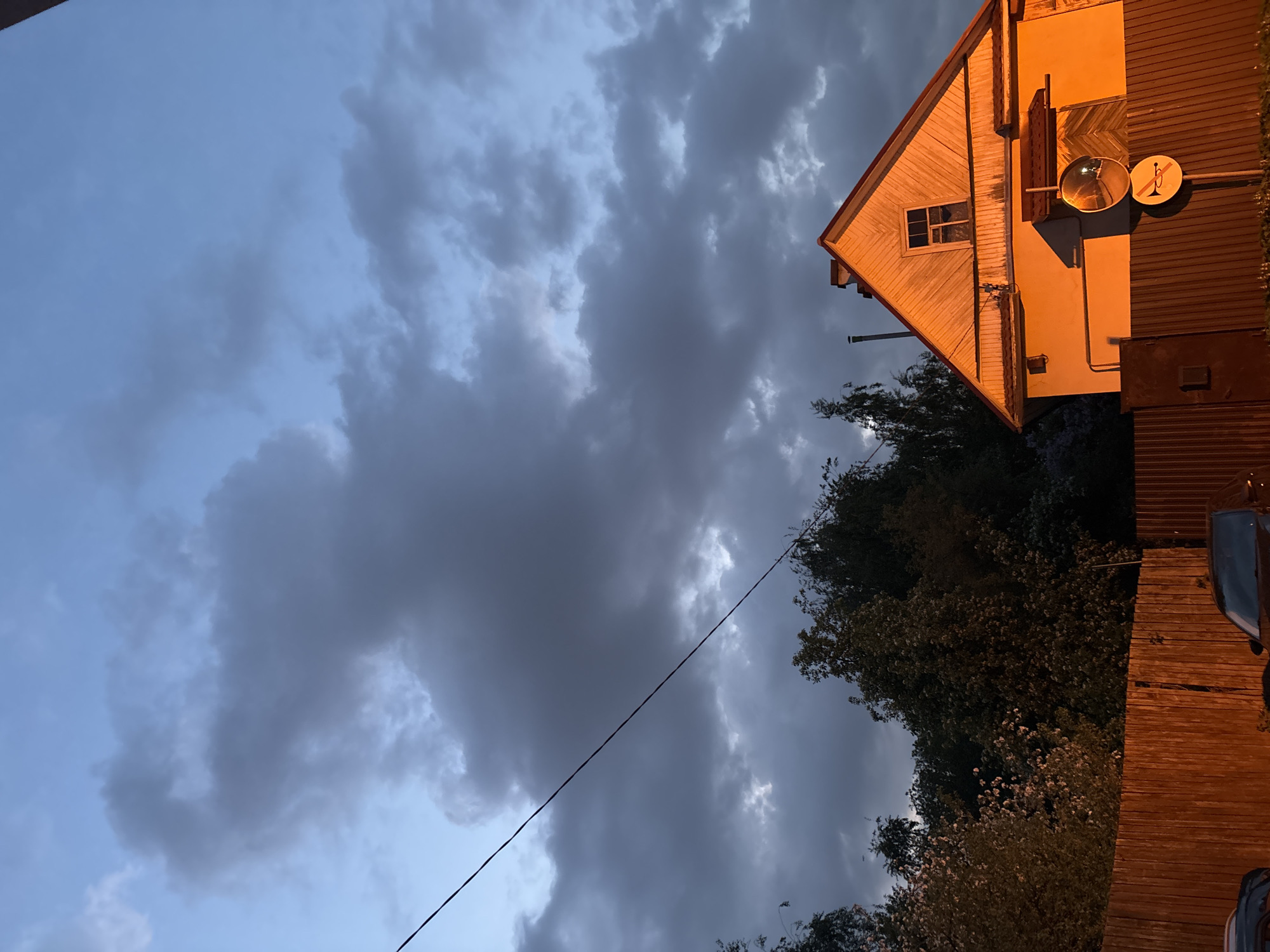 The image shows a view of a residential area in Saint Petersburg, Russia at dusk. A wooden, two-story building with a pitched roof occupies the upper right portion of the frame. The building is illuminated, with a visible window, a satellite dish, and what appears to be a security mirror mounted on its side. Adjacent to the building is a dark, corrugated metal structure. Below the building, foliage obscures the lower part of the frame. A power line stretches diagonally across the frame, from the lower left to the upper right, partially obscuring the sky. The sky is overcast with dark, heavy clouds. Patches of lighter sky are visible around the edges of the cloud cover. The scene appears to be a typical residential area with older buildings and mature trees.