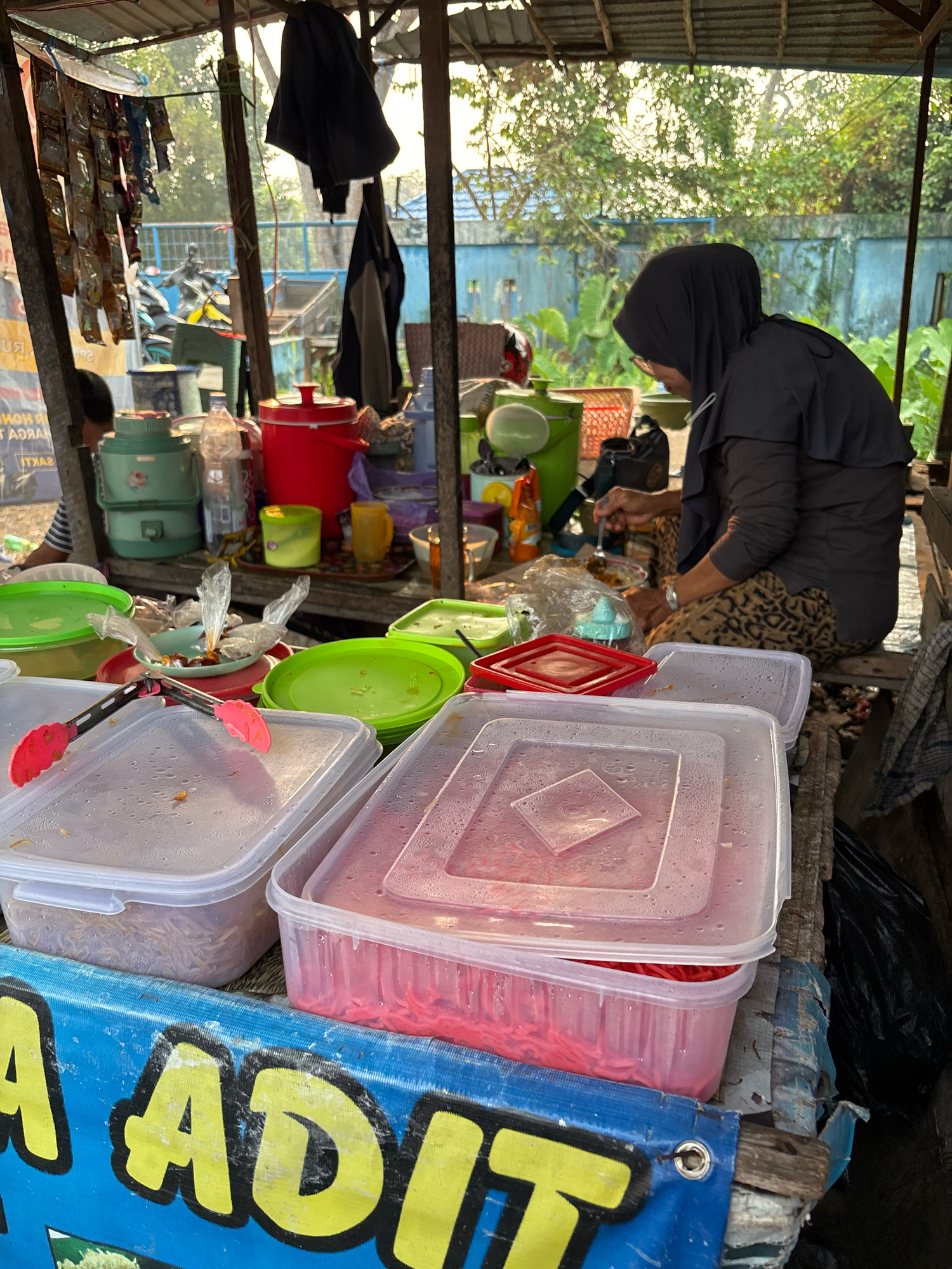 A woman wearing a black hijab and patterned trousers is sitting and preparing food at an outdoor stall. She is using a spoon to mix food in a small bowl. The stall is covered with a corrugated metal roof supported by wooden beams. Various containers, including green and red plastic tubs, and clear plastic food storage containers filled with noodles and other food items, are visible. Red tongs are placed on top of a clear container. A blue banner with yellow lettering that partially reads "ADIT" is in the foreground. The background shows trees and foliage, suggesting a semi-urban or rural setting in Banjarmasin, Indonesia.