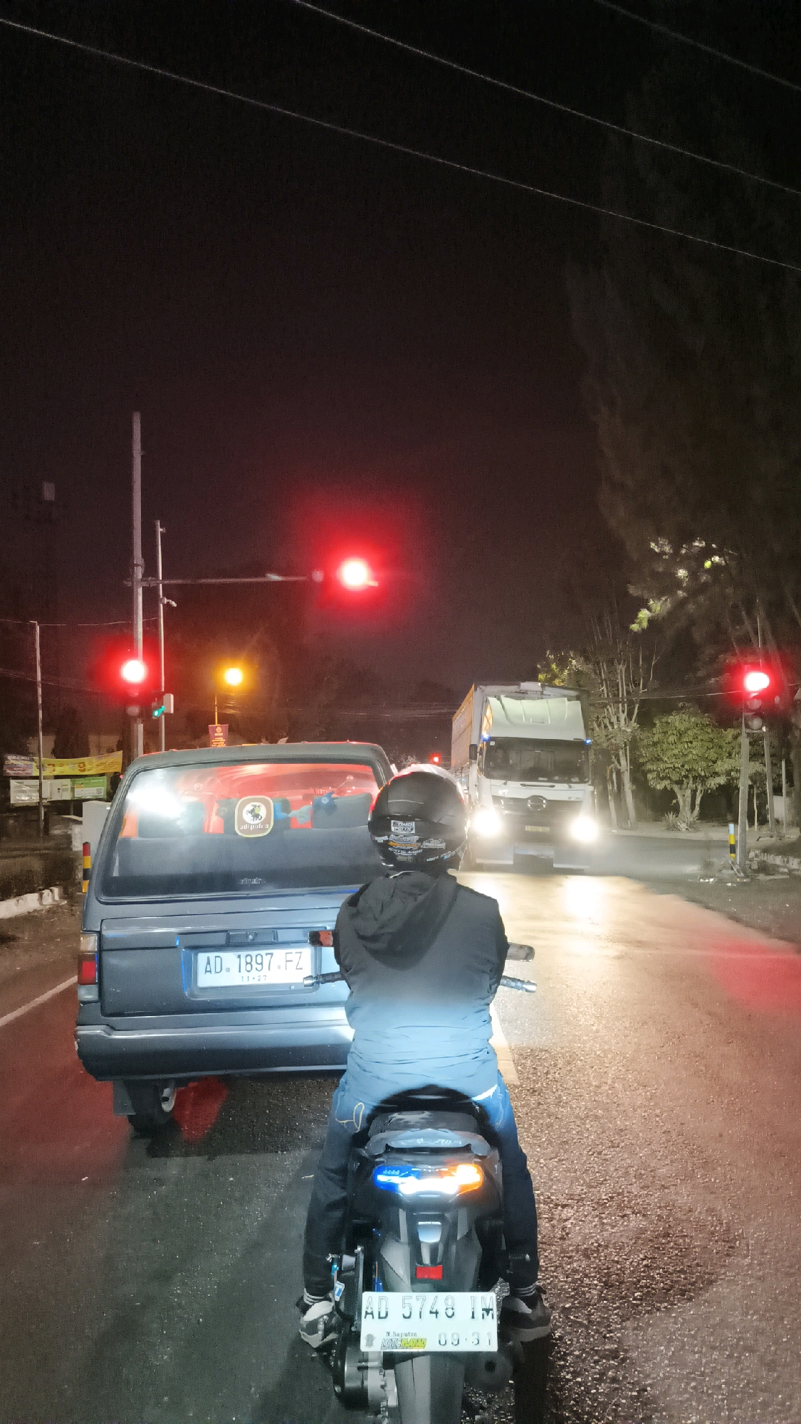 The image depicts a night scene at a traffic light in Drian, Indonesia. A person on a motorcycle, wearing a black helmet and a blue and black jacket, is stopped behind a light blue car. The car has a license plate that reads "AD. 1897. FZ". The motorcycle has a license plate that reads "AD 5748 IM" with the text "Seputra" and "09 31" below it. Red traffic lights are illuminated, indicating that vehicles should stop. A large white truck with its headlights on is visible in the background, approaching the intersection. The road appears wet, suggesting recent rain. Trees and utility poles are present along the side of the road.