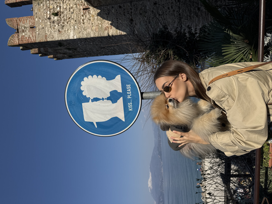 The image depicts an outdoor scene in Sirmione, Italy, specifically near the Scaliger Castle overlooking Lake Garda. In the foreground, a female individual with dark hair, wearing sunglasses and a tan trench coat, is holding a light-brown Pomeranian dog. She is in the act of kissing the dog on its head. The dog is wearing a gray and light-brown knitted garment. Directly behind the subjects, a blue circular sign on a metal pole features two white silhouettes, one with a hat and one with curly hair, facing each other, with the text "KISS... PLEASE" below them. To the right, a large section of a stone and brick castle wall, consistent with the Scaliger Castle's architecture, including crenellated battlements, is visible. In the background, Lake Garda is visible under a clear blue sky, with snow-capped mountains discernible on the distant shore. Sparse vegetation, including leafless trees and green shrubs, populates the midground. The scene is brightly illuminated by direct sunlight.