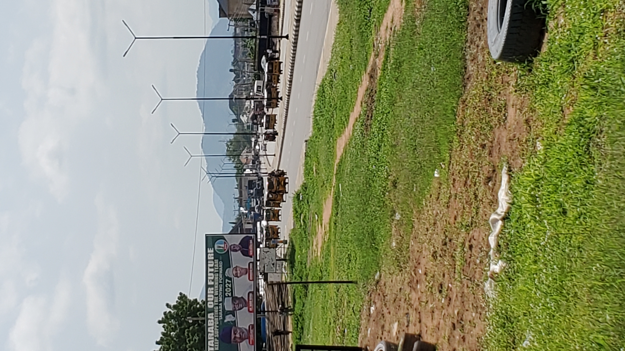 The image depicts a roadside scene in Jalingo, Nigeria. The foreground is dominated by a patch of green grass and a dirt path, with a discarded tire visible on the right side. Beyond this, a road stretches into the distance, lined with a row of streetlights. Along the road, there are several small, three-wheeled vehicles, possibly auto-rickshaws or Keke Napeps, which are common in Nigeria. In the background, hills or mountains can be seen under a cloudy sky.
To the left of the road, a large billboard is prominently displayed. The text on the billboard reads "TARABA OUR FUTURE KEEP SUPPORTING THE WINNING TEAM TARABA MOVING FORWARD 2027". It also features portraits of several men, likely political figures.
The scene suggests a daytime setting, with the lighting indicating it is not early morning or late evening. The weather appears to be partly cloudy. The overall impression is that of a typical urban or semi-urban environment in Nigeria, characterized by local transportation, political messaging, and natural scenery in the background. The image is oriented vertically, suggesting it was taken with the camera held in portrait mode, or that the original image has been rotated.