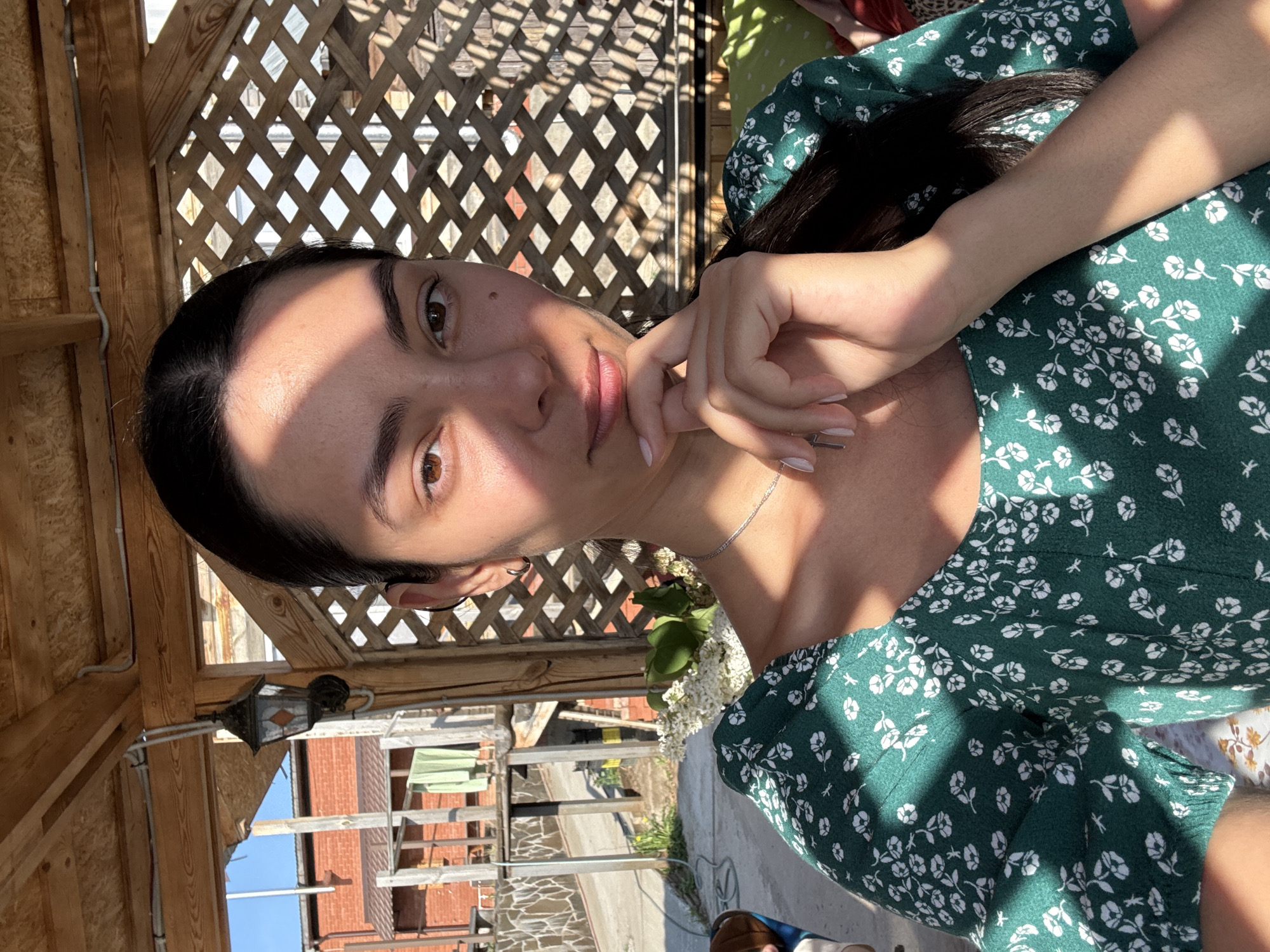 The image depicts a woman with dark hair and brown eyes, wearing a green floral dress. She is indoors, likely in a gazebo or covered patio, based on the wooden lattice structure in the background. Outside the structure, brick buildings are visible, indicating an urban environment. The location is specified as Chaikovsky, Russia. The woman is holding her hand near her face, suggesting a pensive or relaxed pose. There is a light fixture hanging from the wooden structure.