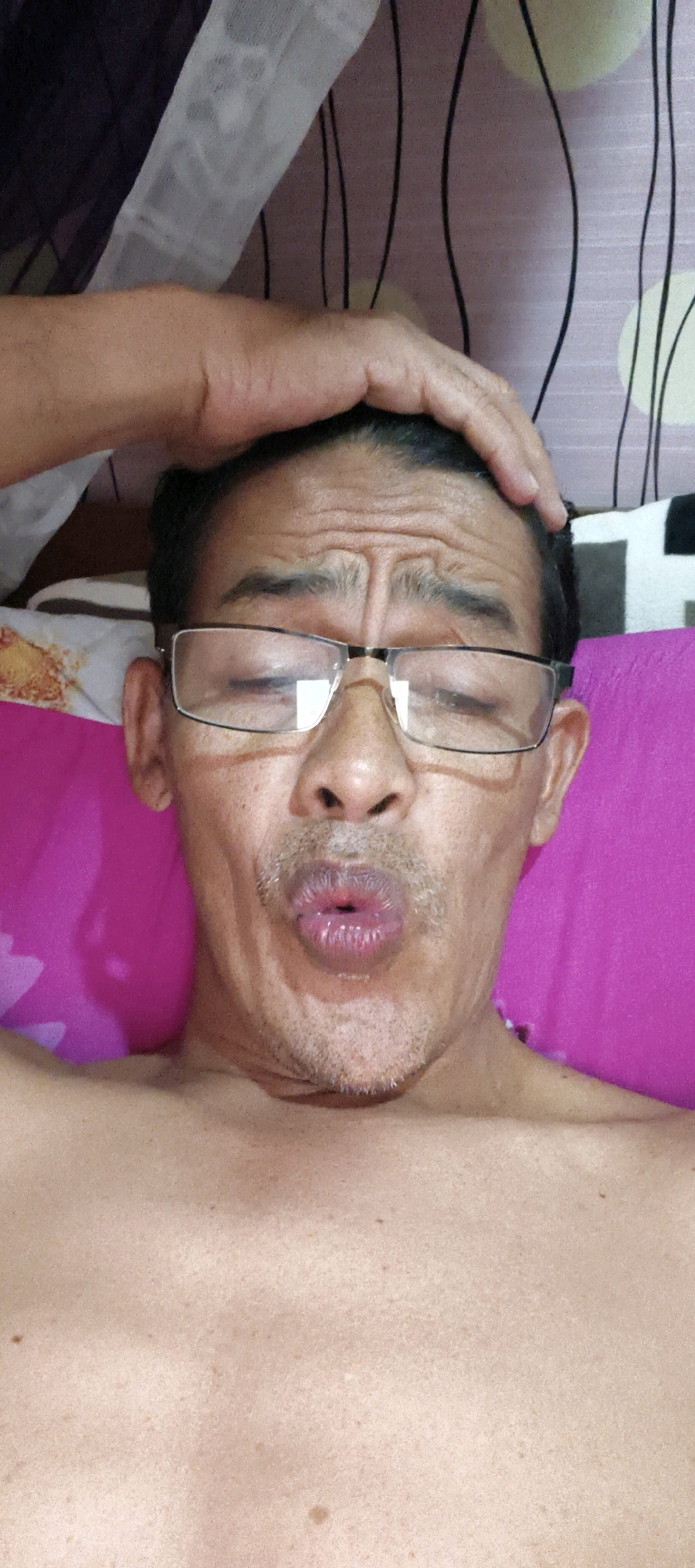 An overhead, close-up shot shows a middle-aged man in Muaro Jambi, Indonesia, making a puckered expression with his lips, as if blowing a kiss. His eyes are closed, and his forehead is wrinkled. He is wearing glasses. His hand is placed on top of his head, with his fingers spread out. The background consists of a pink surface, possibly a bed, with a purple and white patterned fabric visible above. The man's chest is bare.