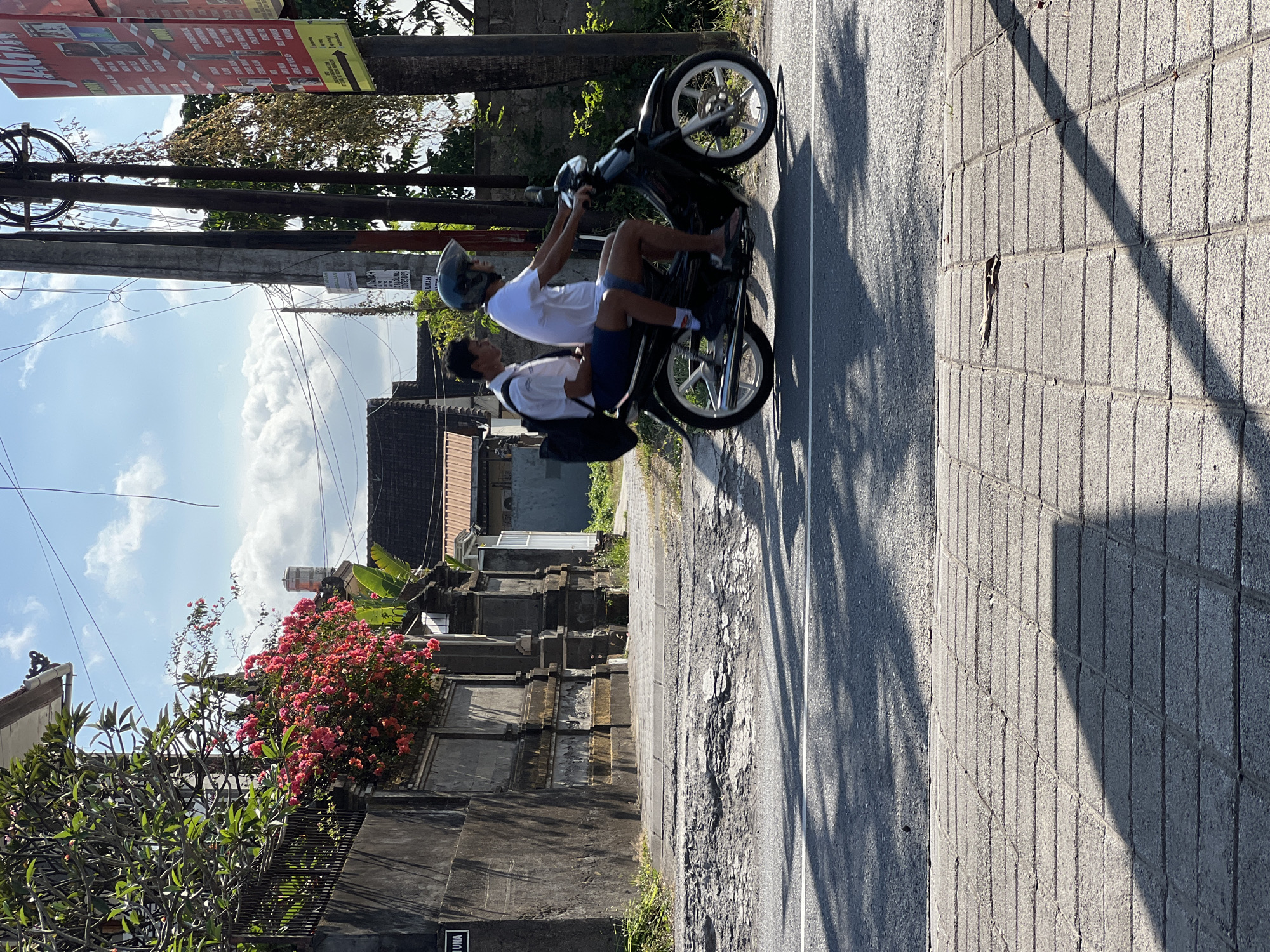 Two young males are riding a black motorcycle on a paved road in Denpasar, Indonesia. The rider in front wears a white t-shirt and blue shorts, while the passenger behind him wears a white collared shirt and blue shorts. Both are wearing helmets. The motorcycle has a silver exhaust pipe and white-rimmed wheels. The road has a white line painted on it. To the left of the road is a grey stone wall with a dark iron fence on top, and vibrant pink bougainvillea flowers cascading over it. Behind the wall, a traditional Balinese-style building with a red-tiled roof is visible. To the right of the road, there are utility poles with numerous wires, and a red advertisement sign. The environment appears sunny with scattered clouds in a blue sky, and the ground is paved with grey rectangular bricks. The scene suggests a typical day in a Balinese urban or suburban setting.