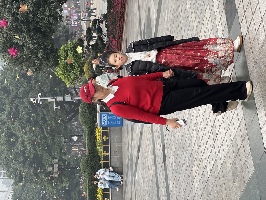 An older woman in a red sweater and cap holds hands with a young girl wearing a floral-patterned dress and black jacket on a paved walkway. The woman holds a white object in her other hand. A lush green area with trees and small bushes frames the background, with some yellow flowers in the middle ground. People are visible in the distant background. Small yellow bird-like shapes appear to be floating in the air.
