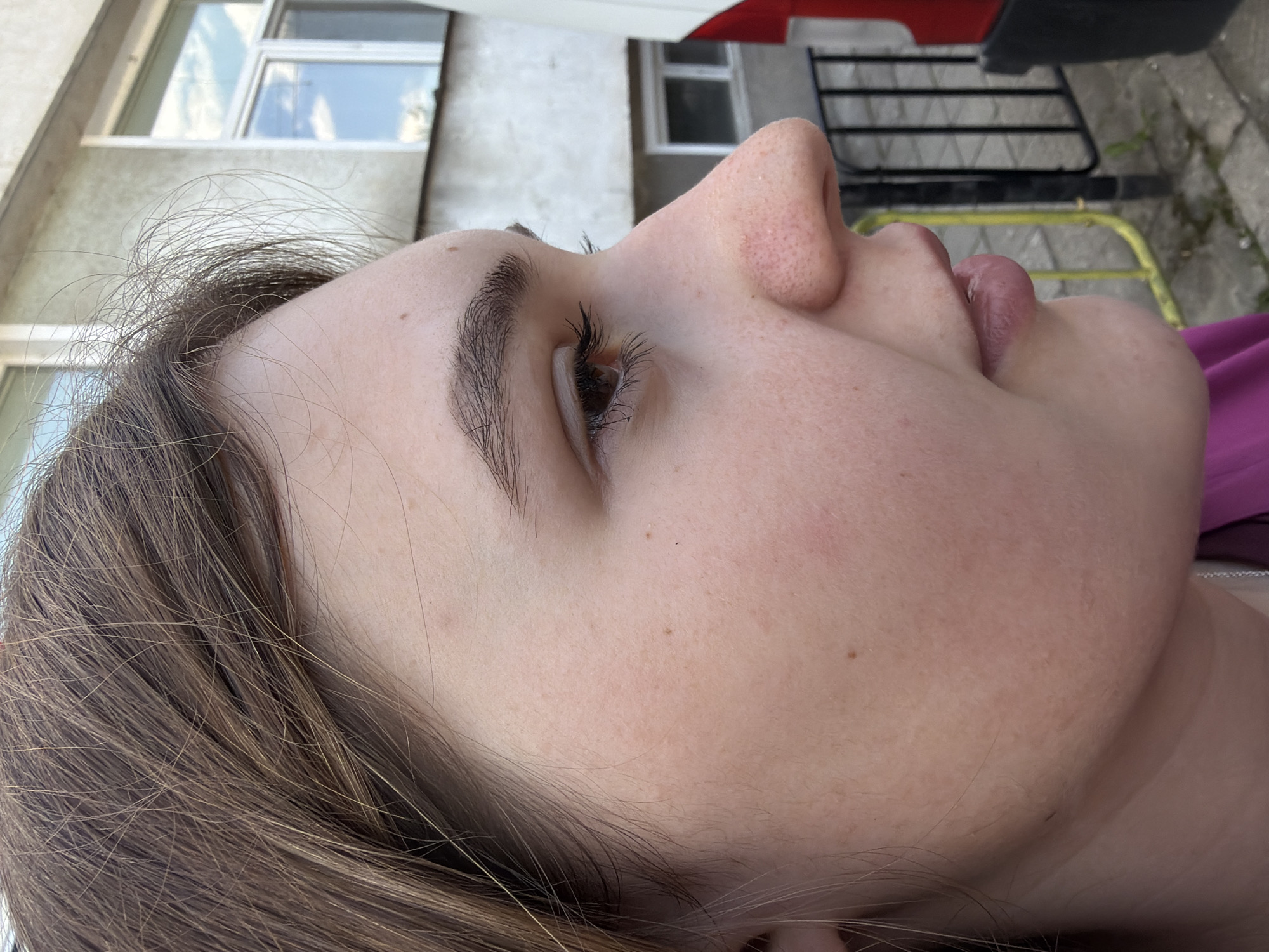 The image shows a close-up, side profile of a person's face. The person has light skin, brown hair, and visible eyelashes. The nose is prominent and slightly upturned. The lips are partially open. The background includes a building with windows, a section of a vehicle (possibly a car or bus) with red and white paint, a metal structure (possibly a fence or gate), and pavement. Based on the metadata location (Panagyurishte, Bulgaria), the scene is likely located within that city.