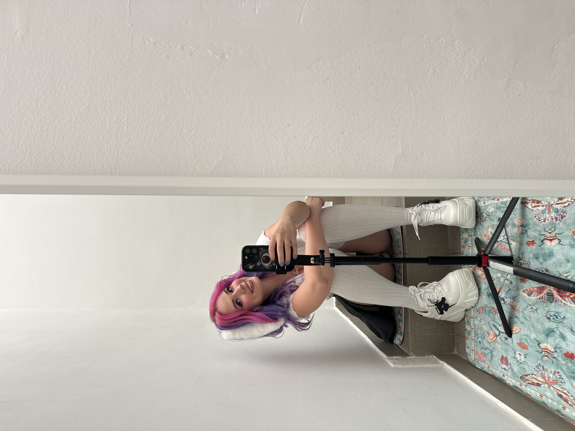 A young woman with pink and purple hair, wearing white knee-high socks and chunky white sneakers, is seated on the floor. She is holding a smartphone attached to a small tripod and appears to be taking a selfie. The room has a white wall on the left and a patterned rug on the floor. The location is inferred as City of Zagreb, Croatia, based on provided location data.